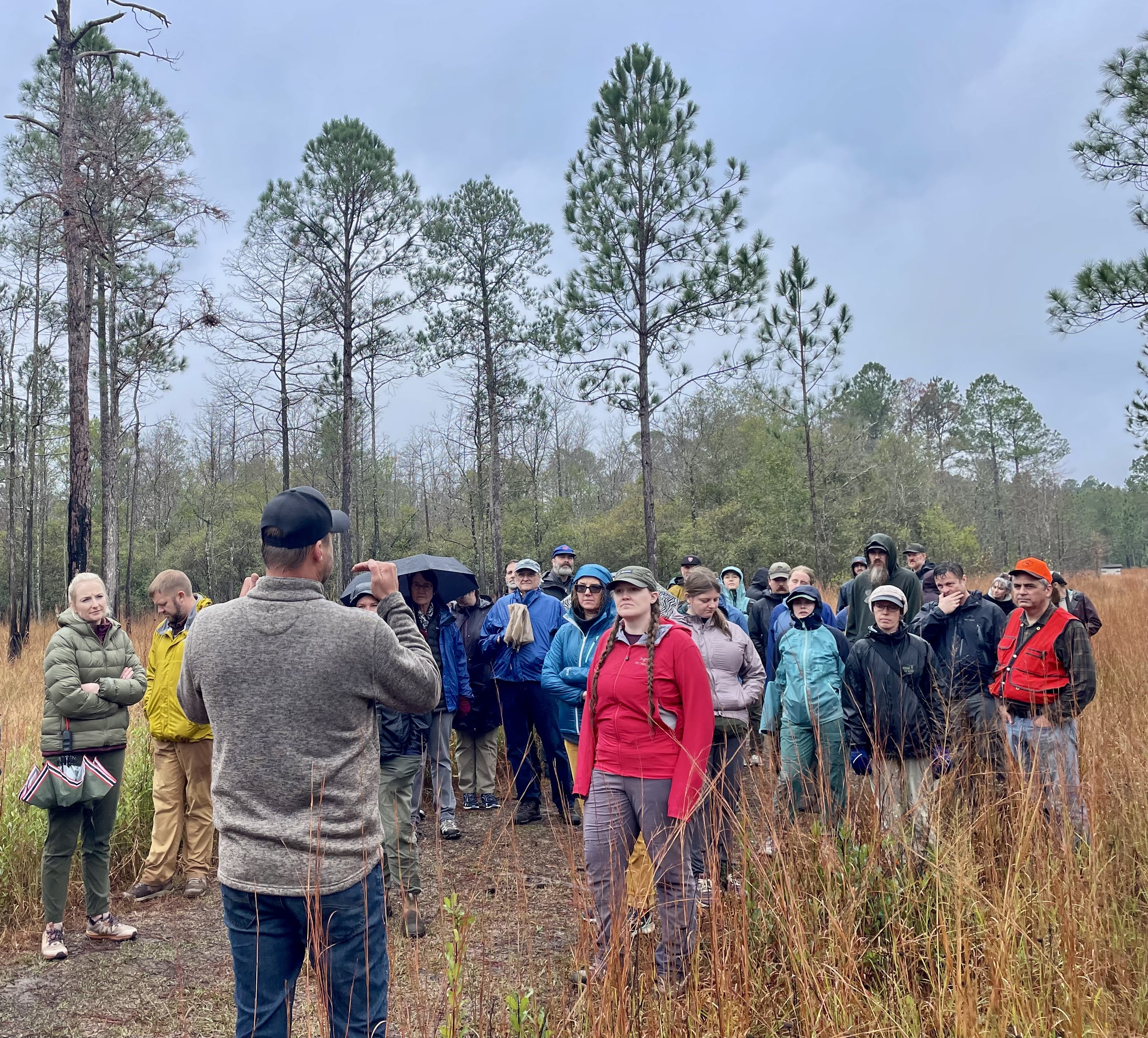 A group of people wearing rain gear gather to listen to a talk about the surrounding forest.