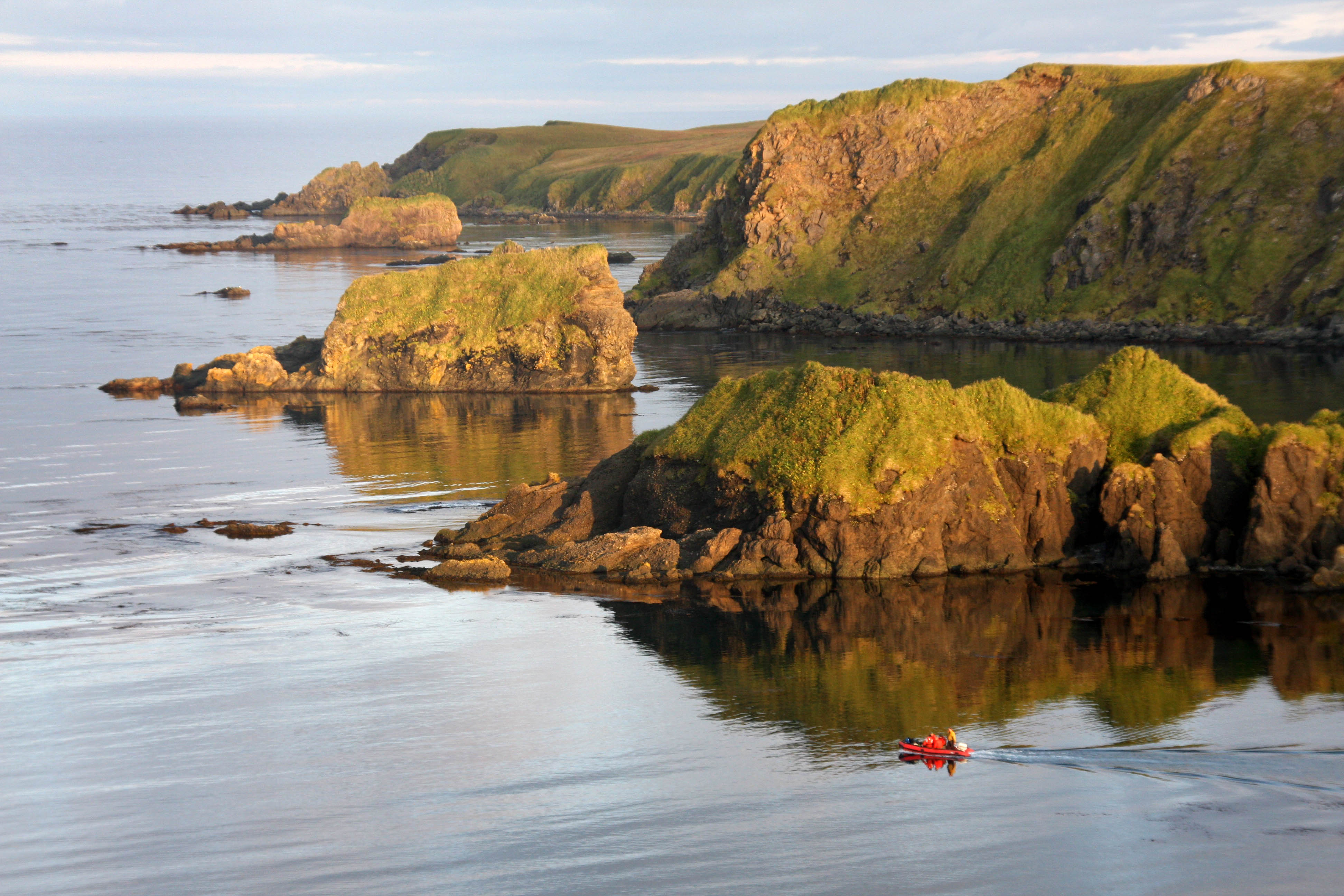 A few people in a zodiac boat travel past the large rocky Aleutian Islands.