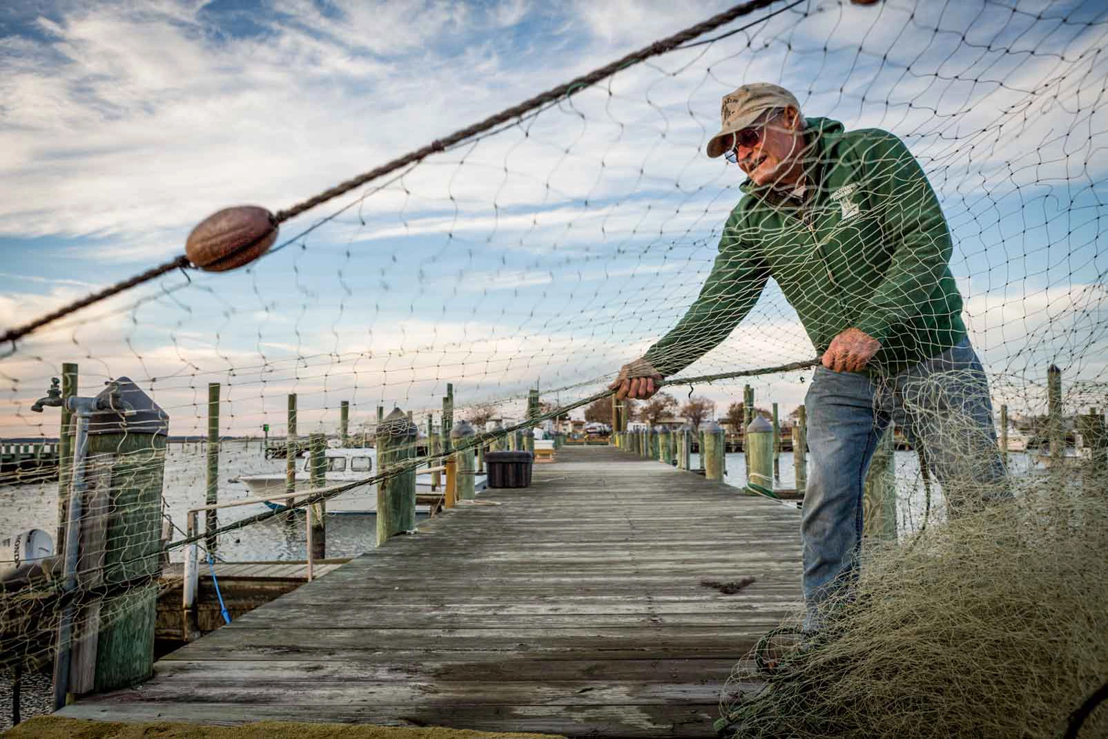 A man stands on a wooden pier pulling in and stacking a net. Small fishing boats are tied up to the pier in the background.