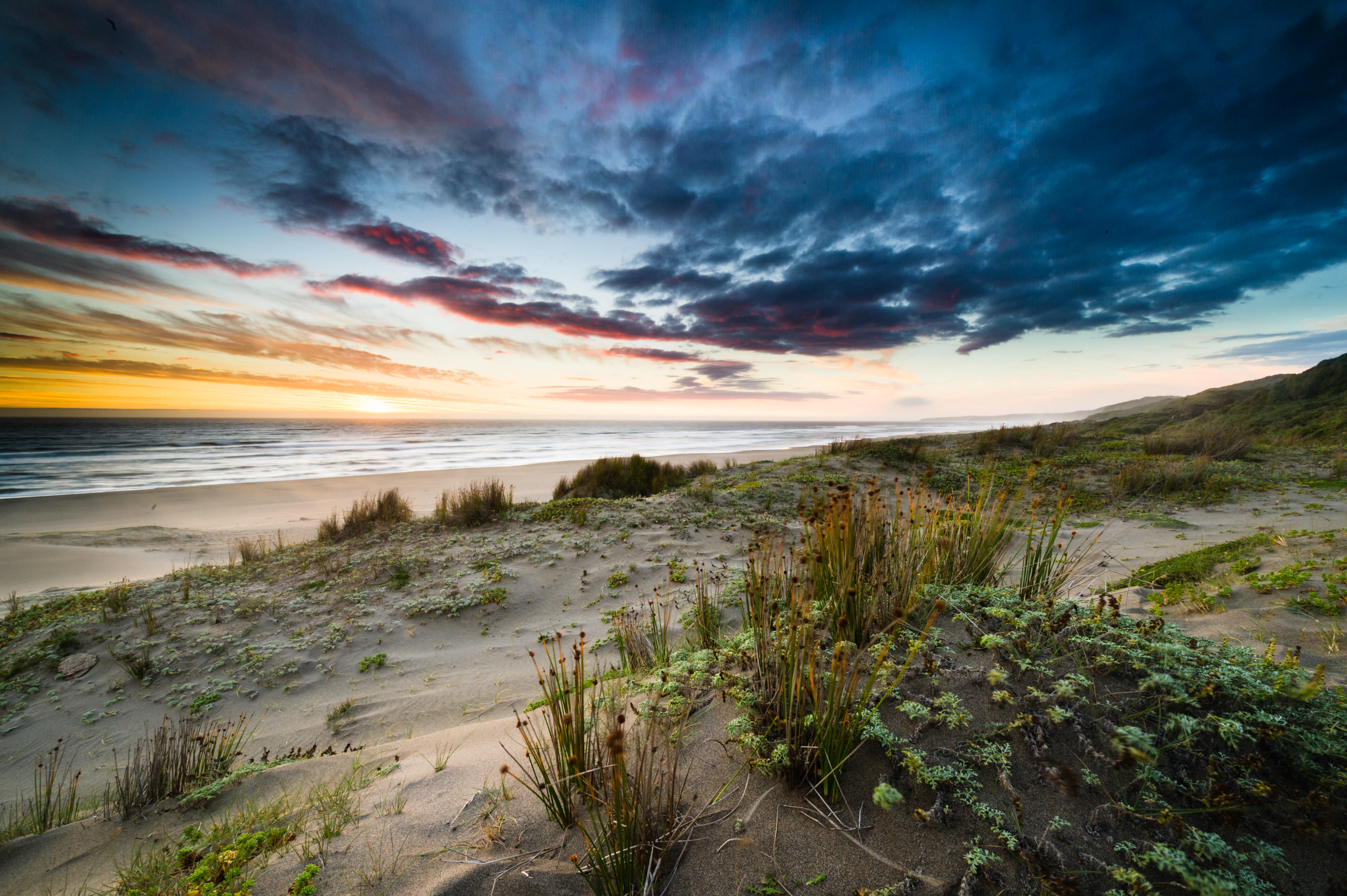 Dramatic landscape view of sand dunes overlooking a beach and ocean under a cloud-filled sky colored in blue and orange.