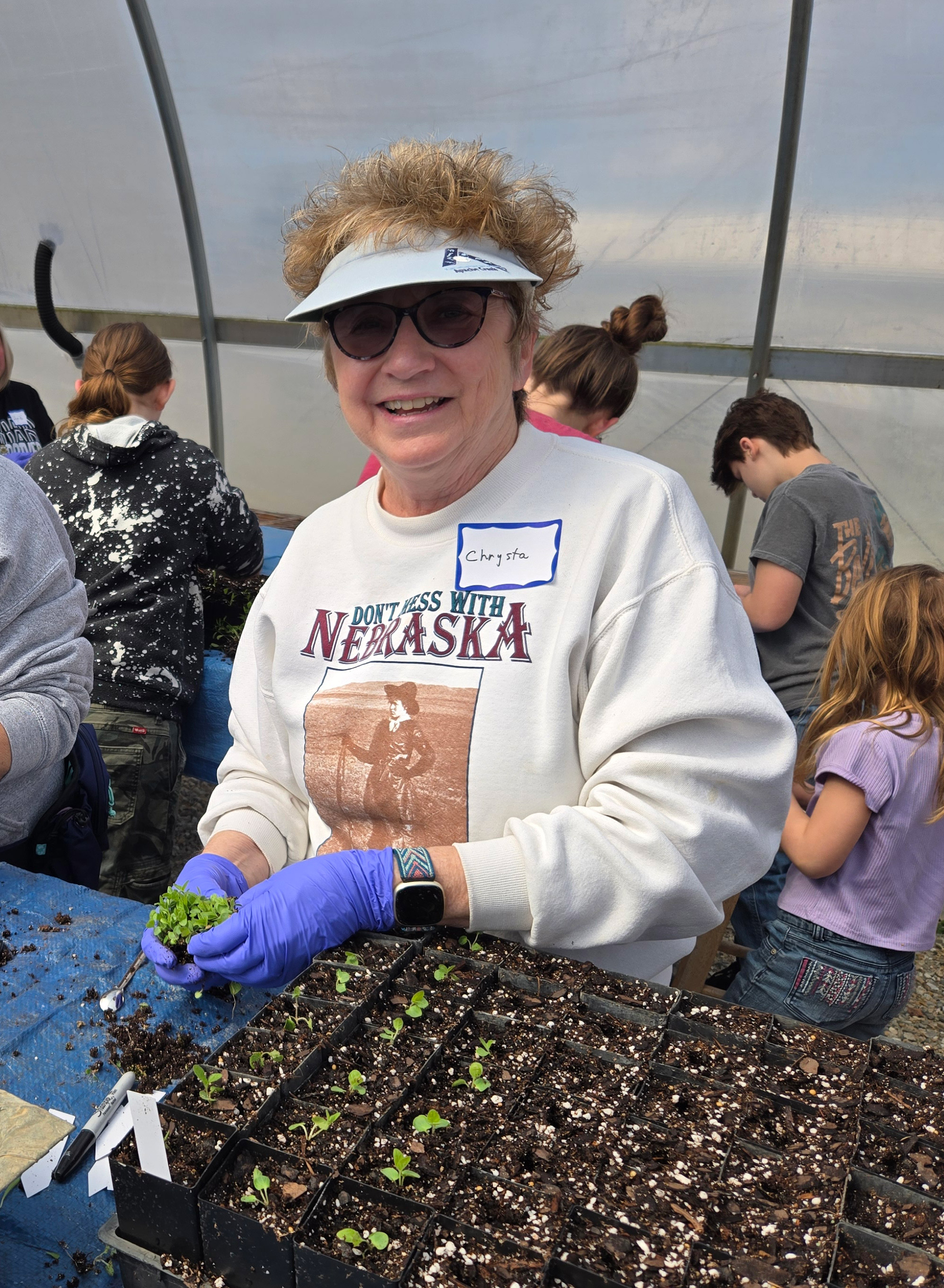 A woman wearing a visor and sunglasses holds seedlings in hands covered with blue gloves. 
