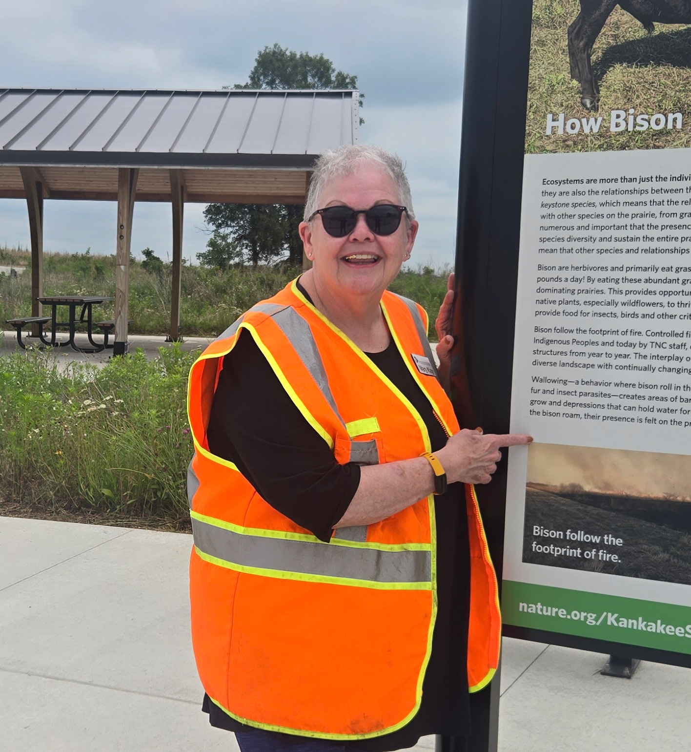 A woman wearing sunglasses and a orange safety vest stands next to an informational sign. 