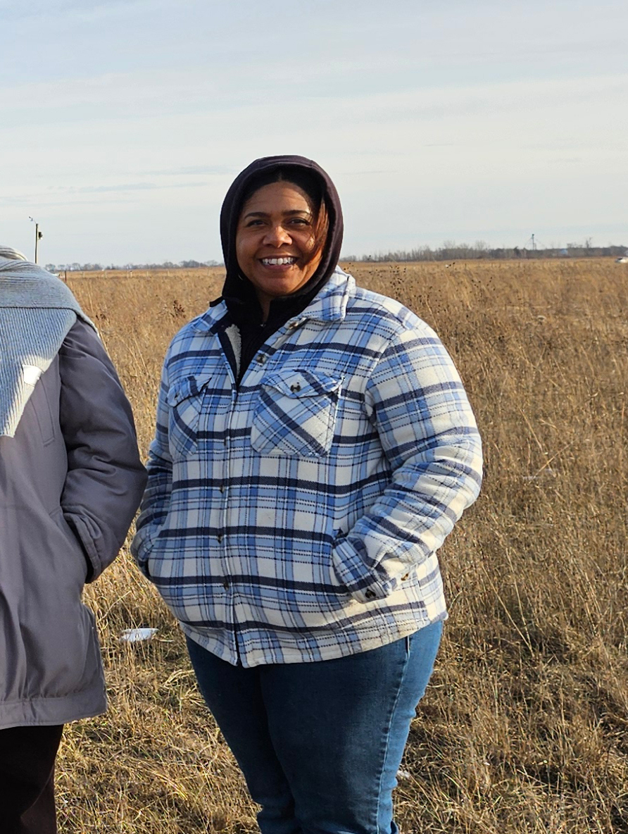 Woman wearing a blue plaid flannel shirt and black hood stands in front of a prairie.