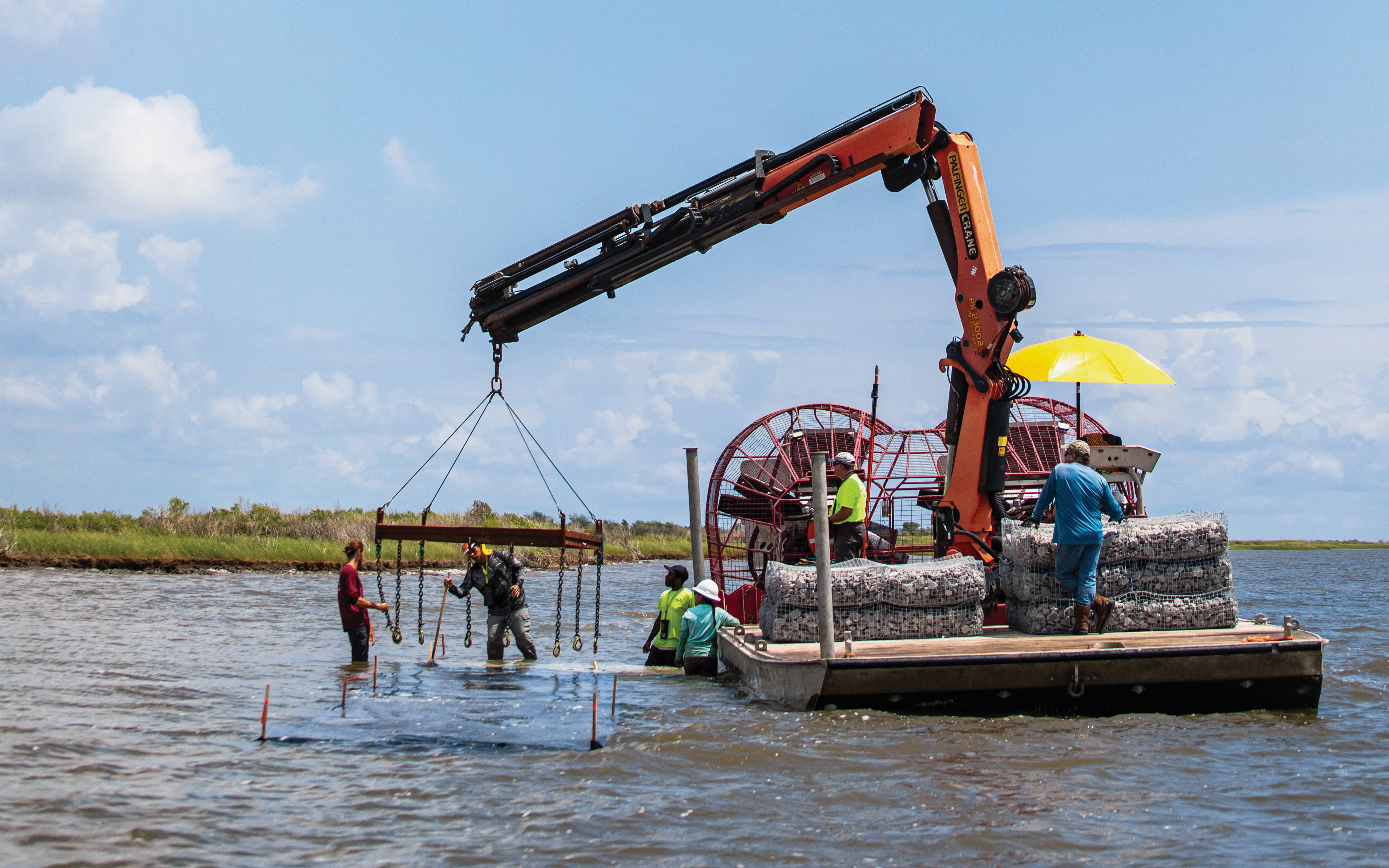 Several people help a flat bottomed boat with a crane through the water.