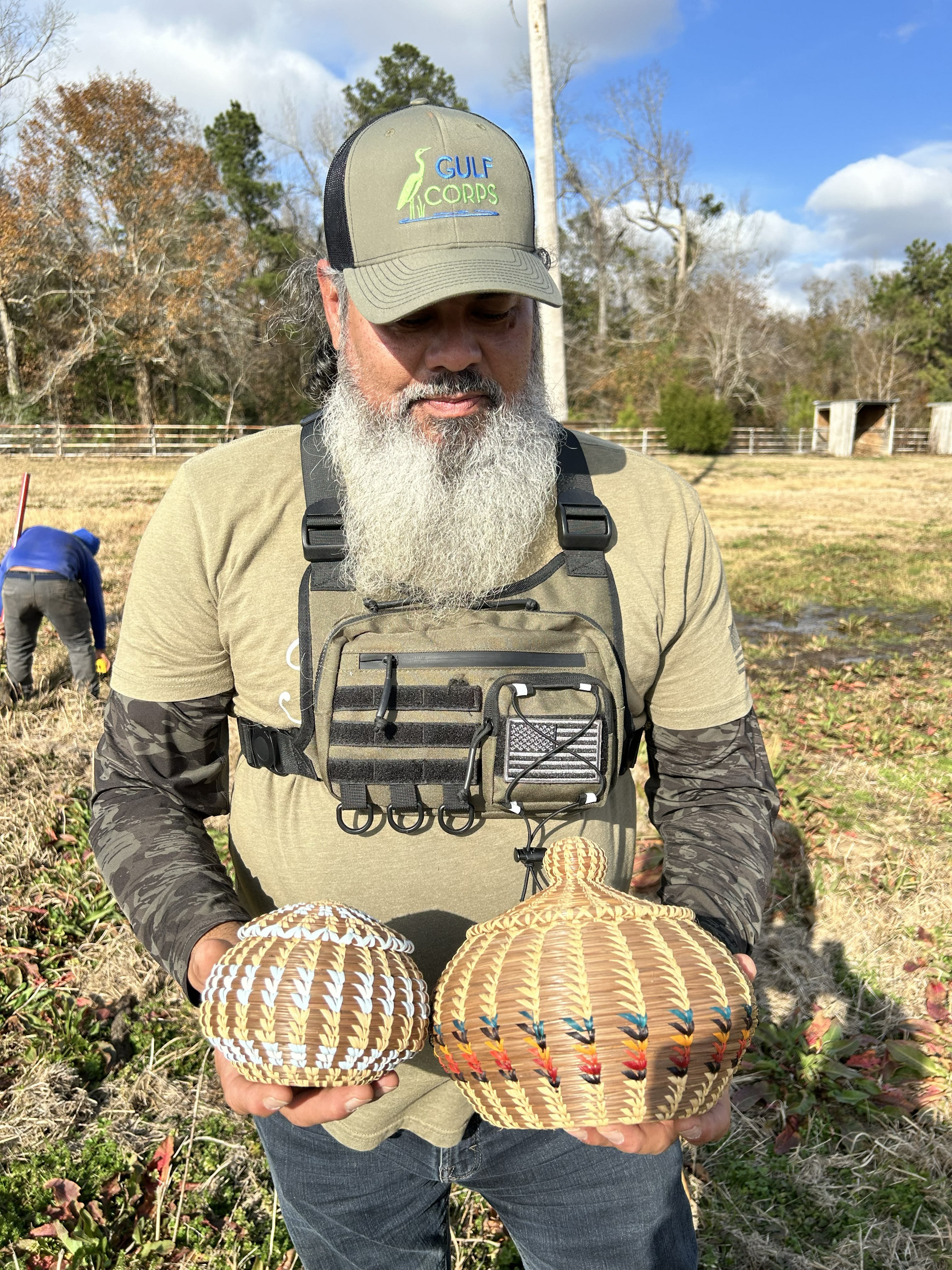 A man wearing a hat holds handmade baskets in each hand. 
