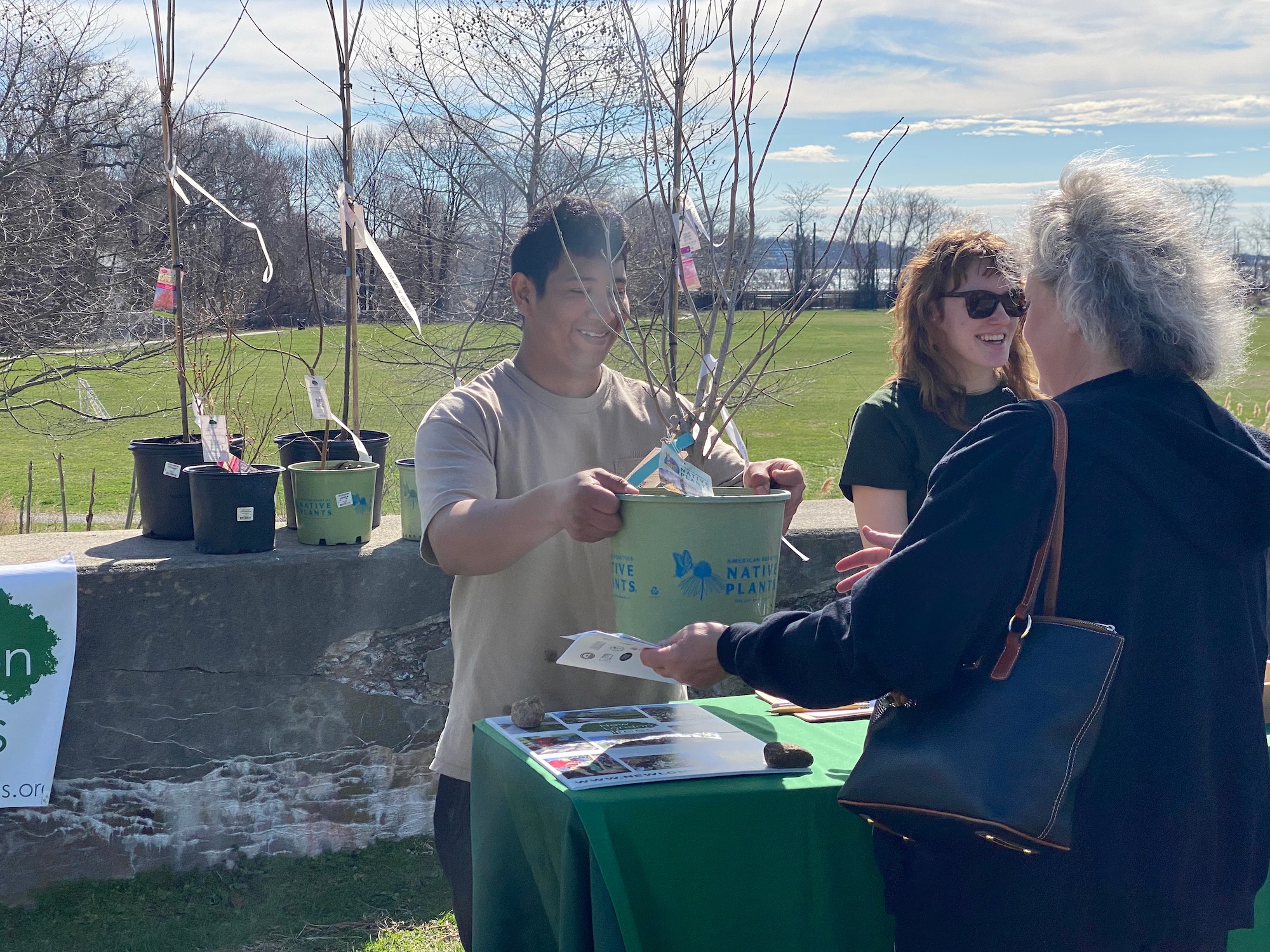 A person is handing a potted plant to another person at an outdoor event. Nearby, someone is smiling. A table with informational materials and a sign that partially reads "Native Plants" is visible. The background features a grassy area and bare trees under a clear sky.