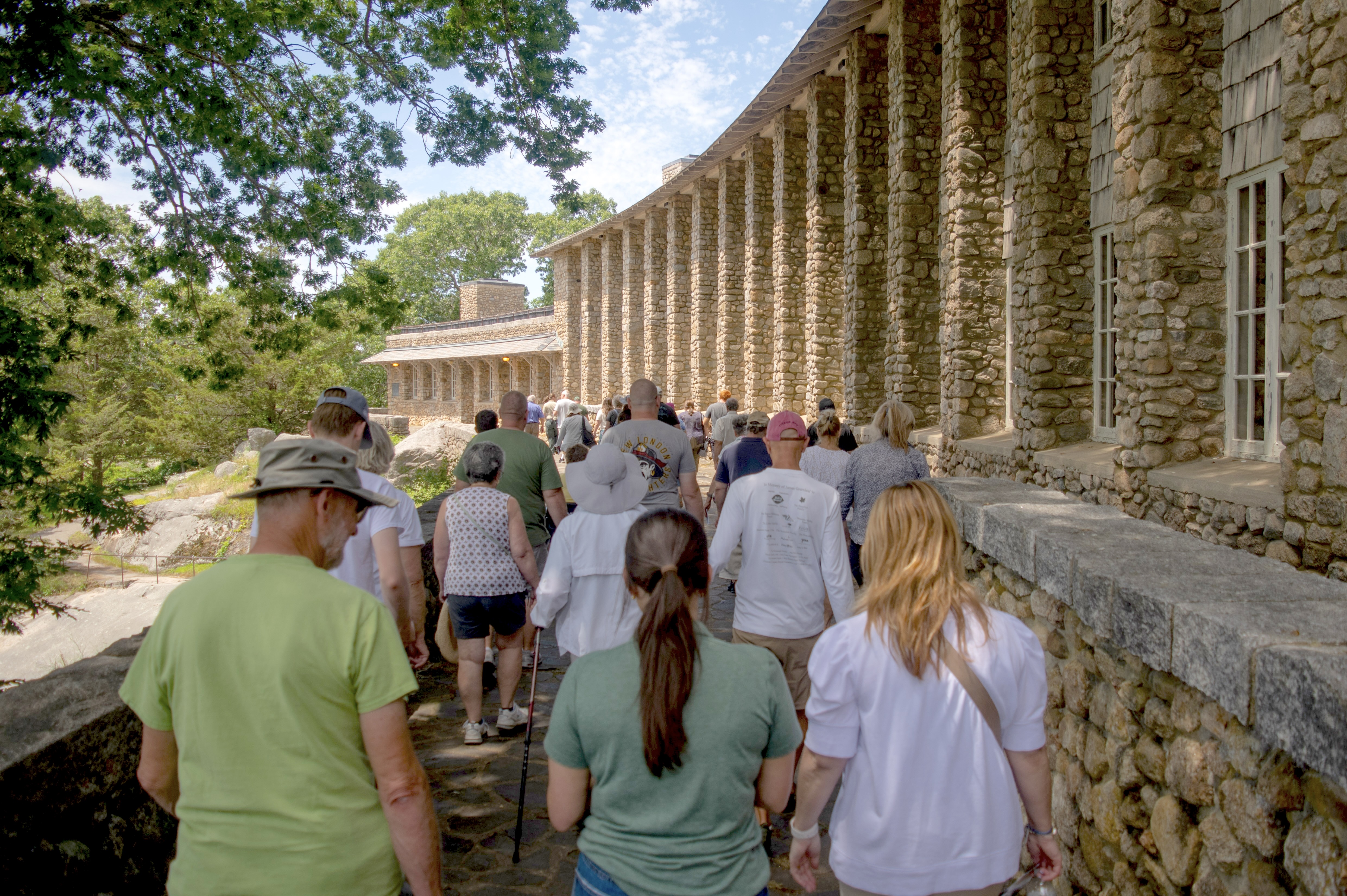 A crowd of people walking along a stone pathway beside the Ellie Mitchell Pavilion at Rocky Neck State Park. The structure features a series of stone columns. Lush greenery surrounds the area with a blue sky overhead.