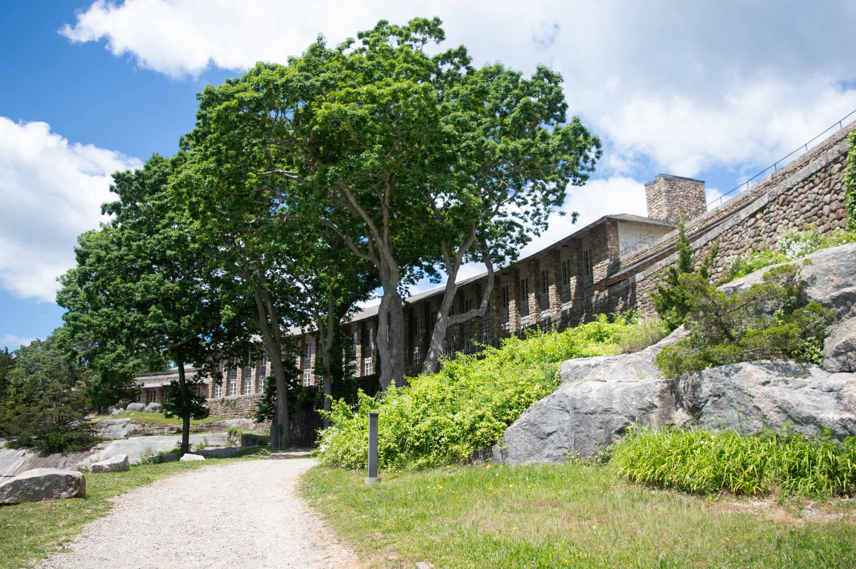 A large stone paviliion at the Rocky Neck State Park is partially obscured by lush green trees. A gravel path winds through well-maintained grass and vegetation under a bright blue sky with scattered clouds.