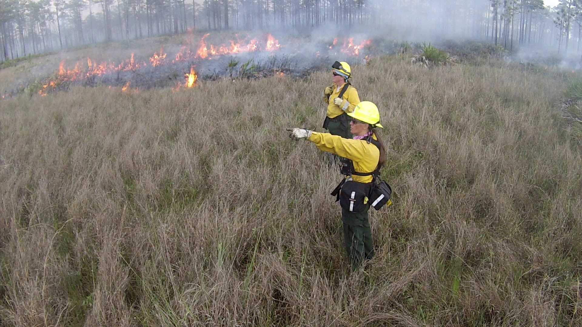 Two women stand in a grassy field wearing fire gear with flames in the background.
