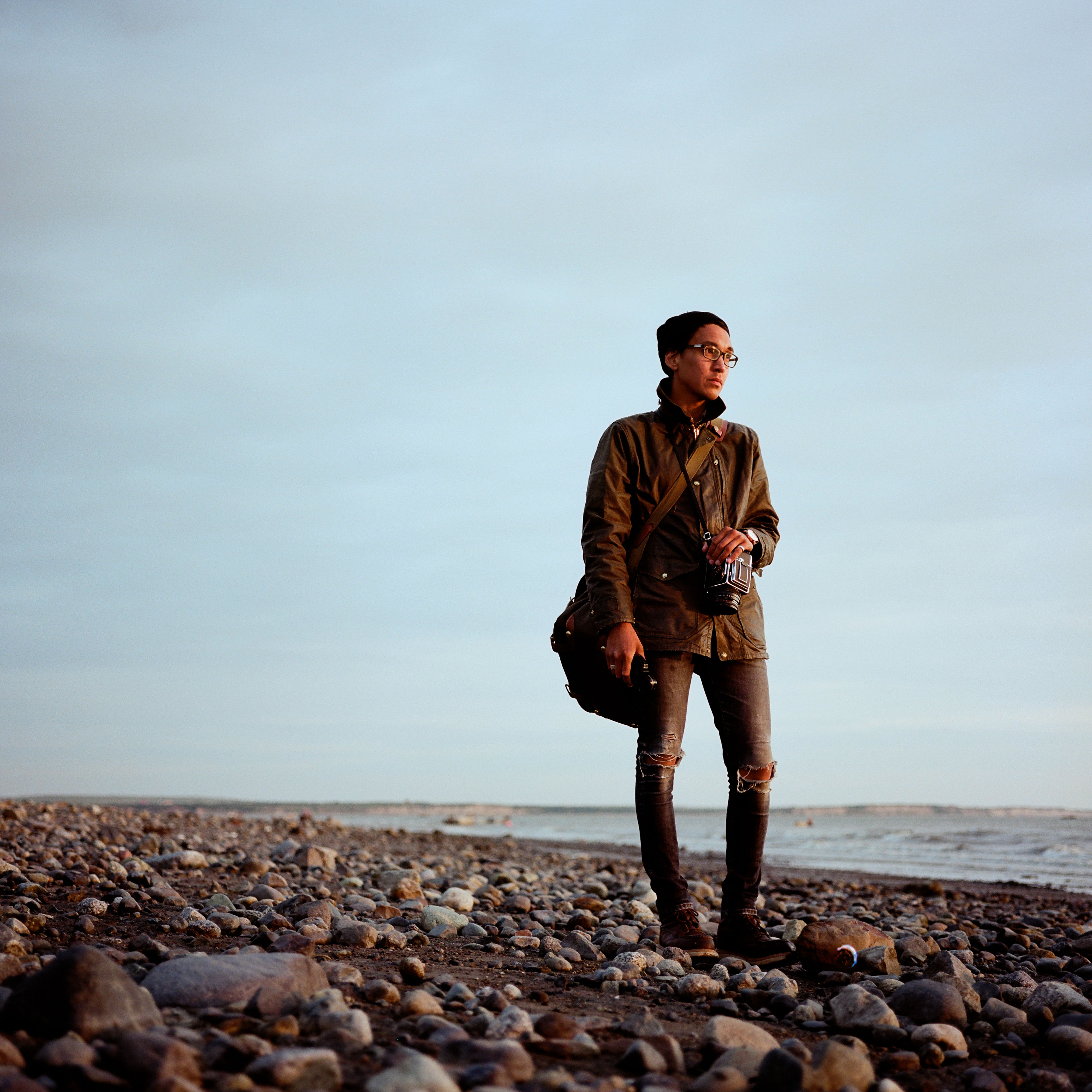 man stands on rocky shore with camera looks away in the distance