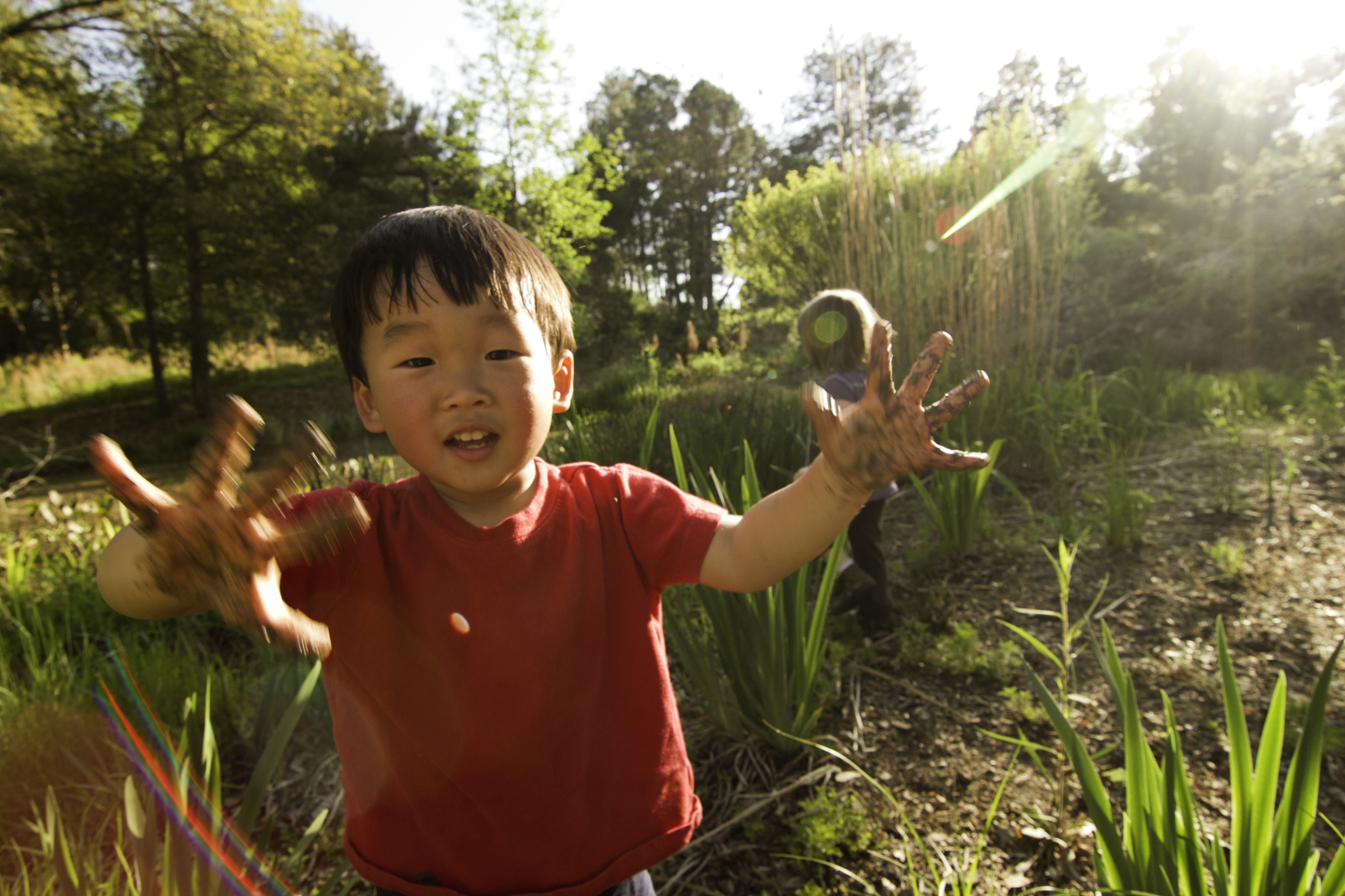 smiling toddler stands in a green space with mud on his hands