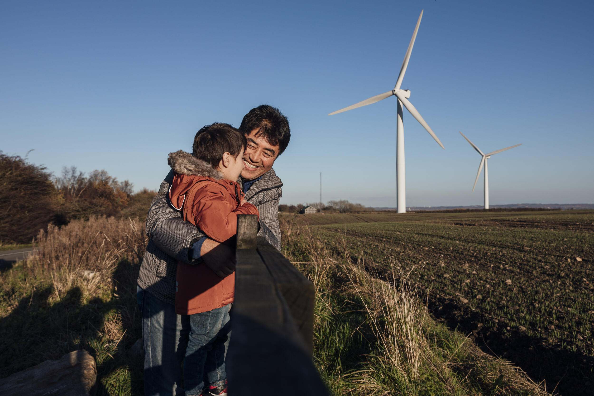 An adult and a child standing outside with a wind turbine in the background. 