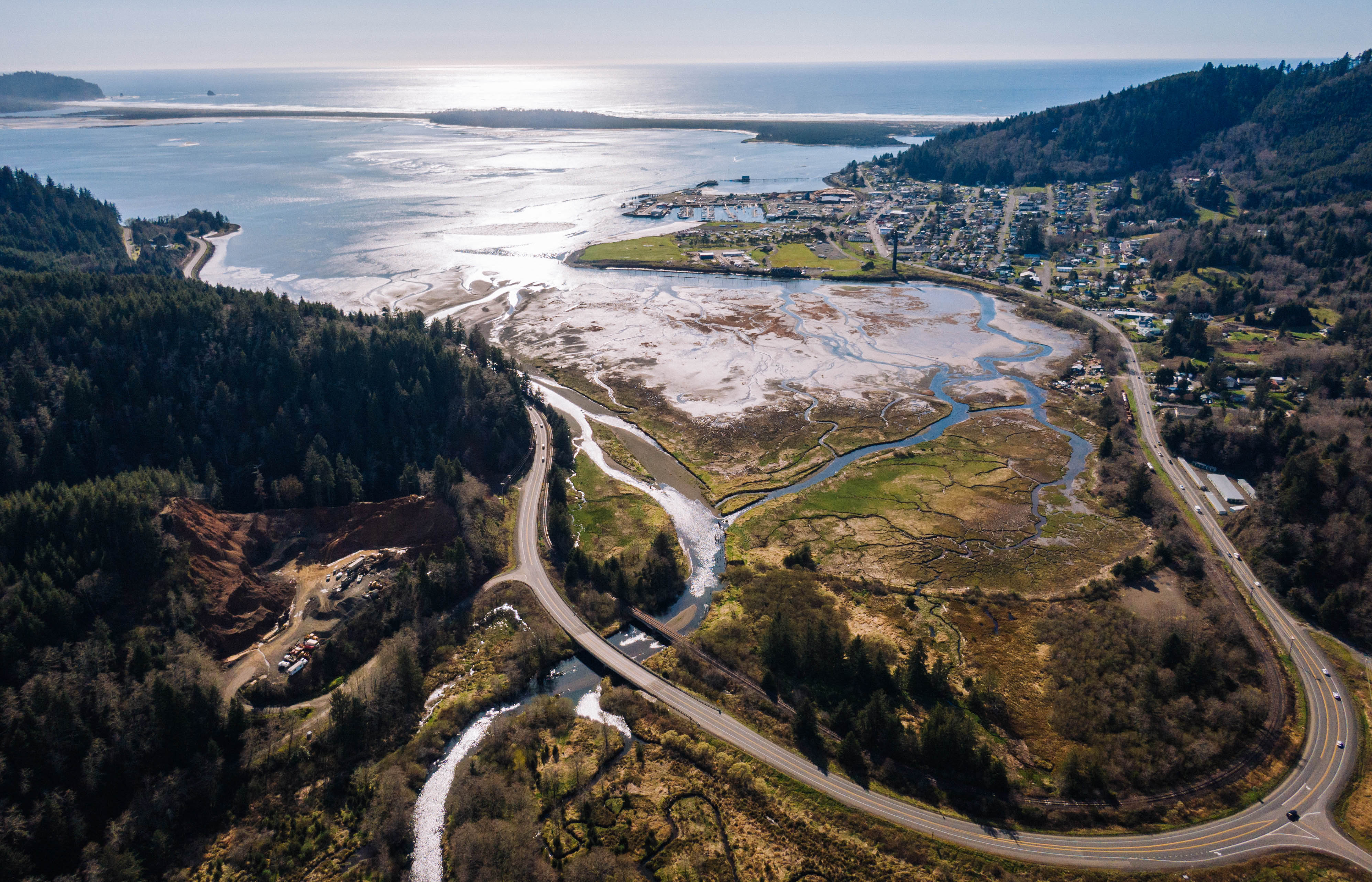 Aerial view of an estuary with meandering and interconnecting streams flowing into a coastal bay, between a town and forests, all along a coastal highway.