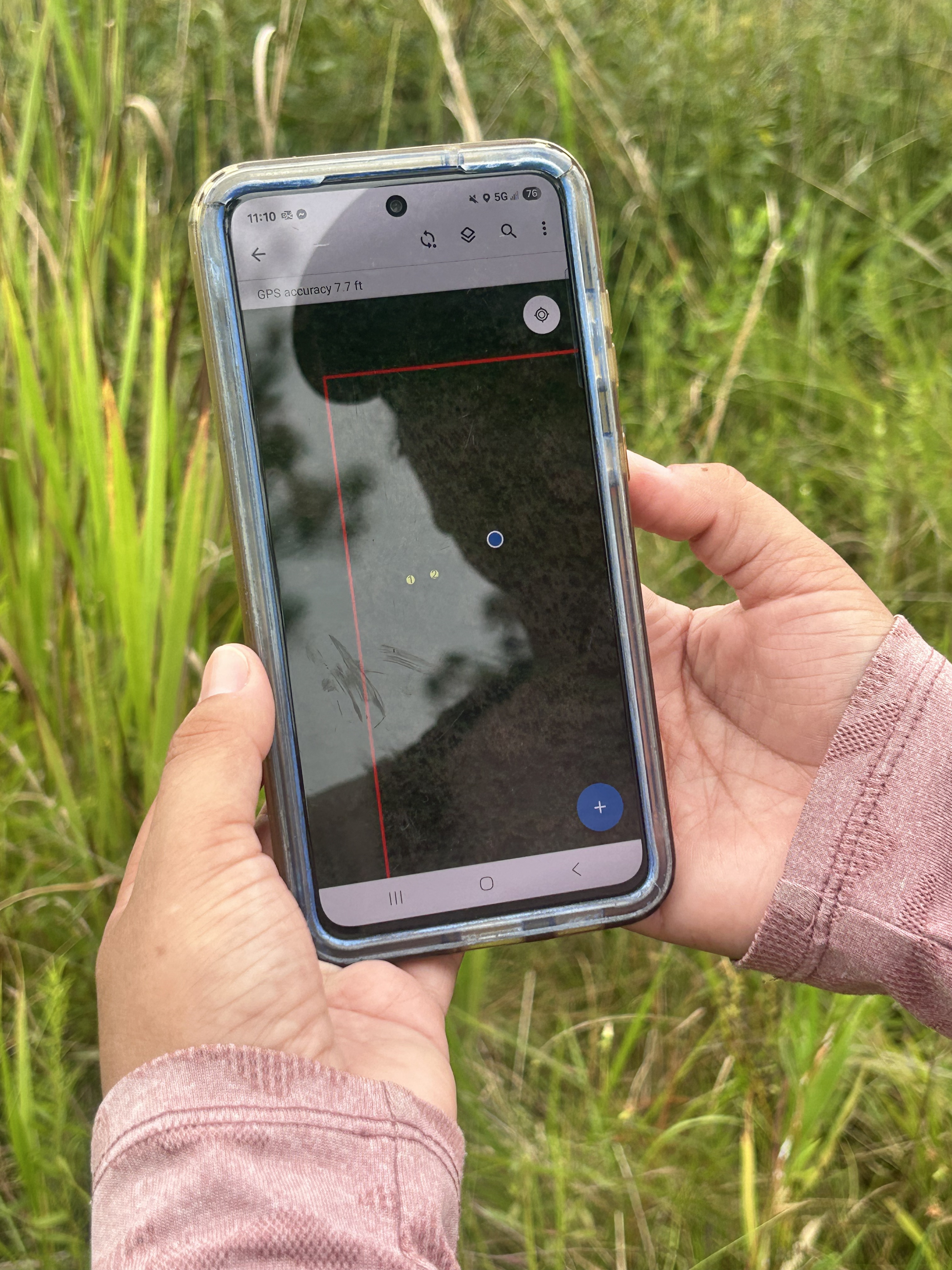 Two hands hod a cell phone over a grassy field.