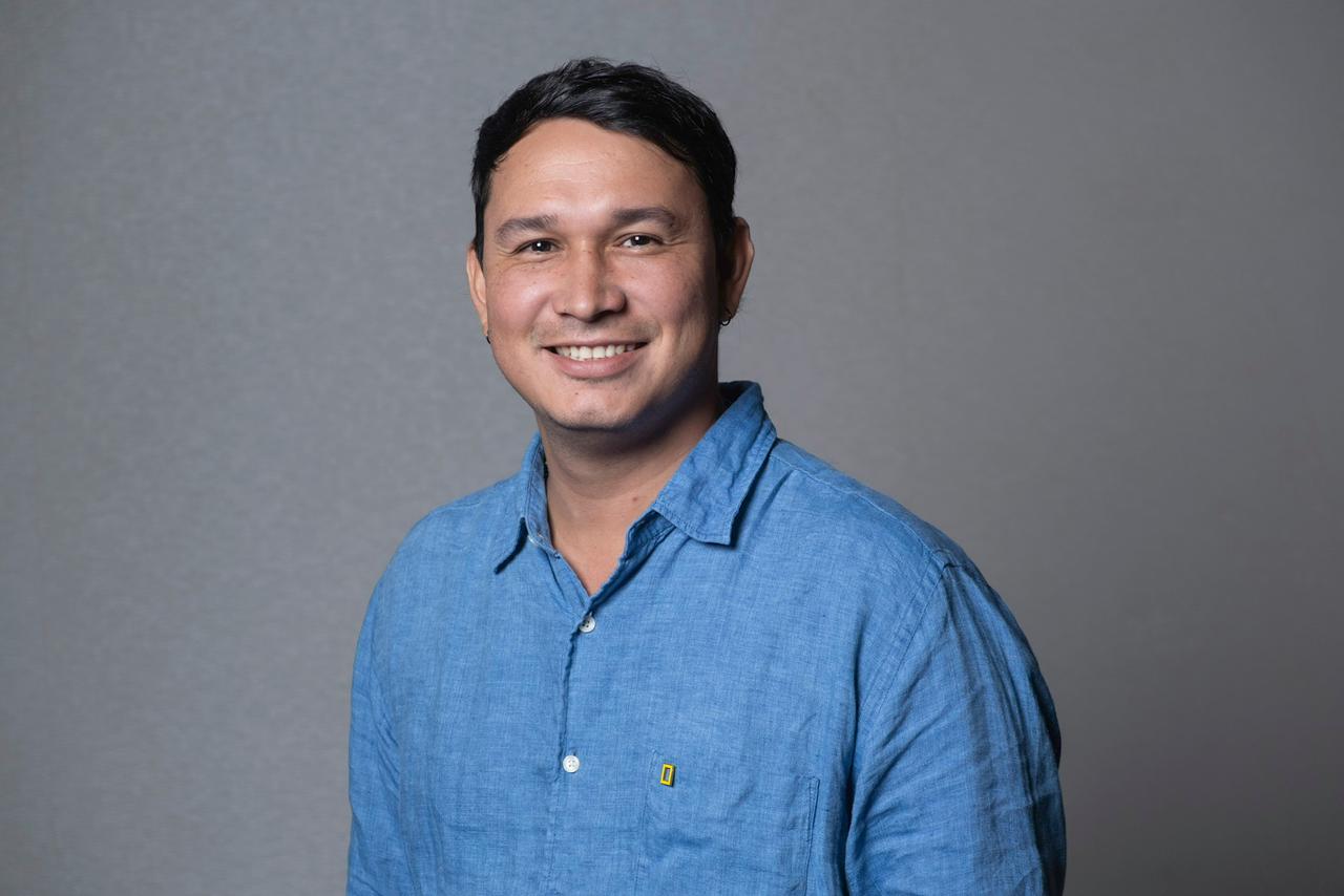 Portrait of Andrés Cardona, wearing a light blue shirt, smiling at the camera, with a grey background. 