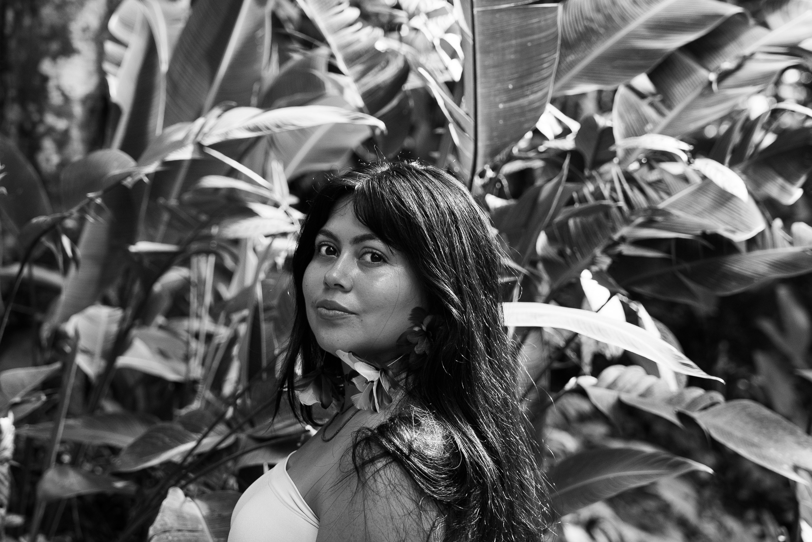 Black and white portrait of Eli Virkina, giving a side look to the camera, surrounded by big palm leaves. 