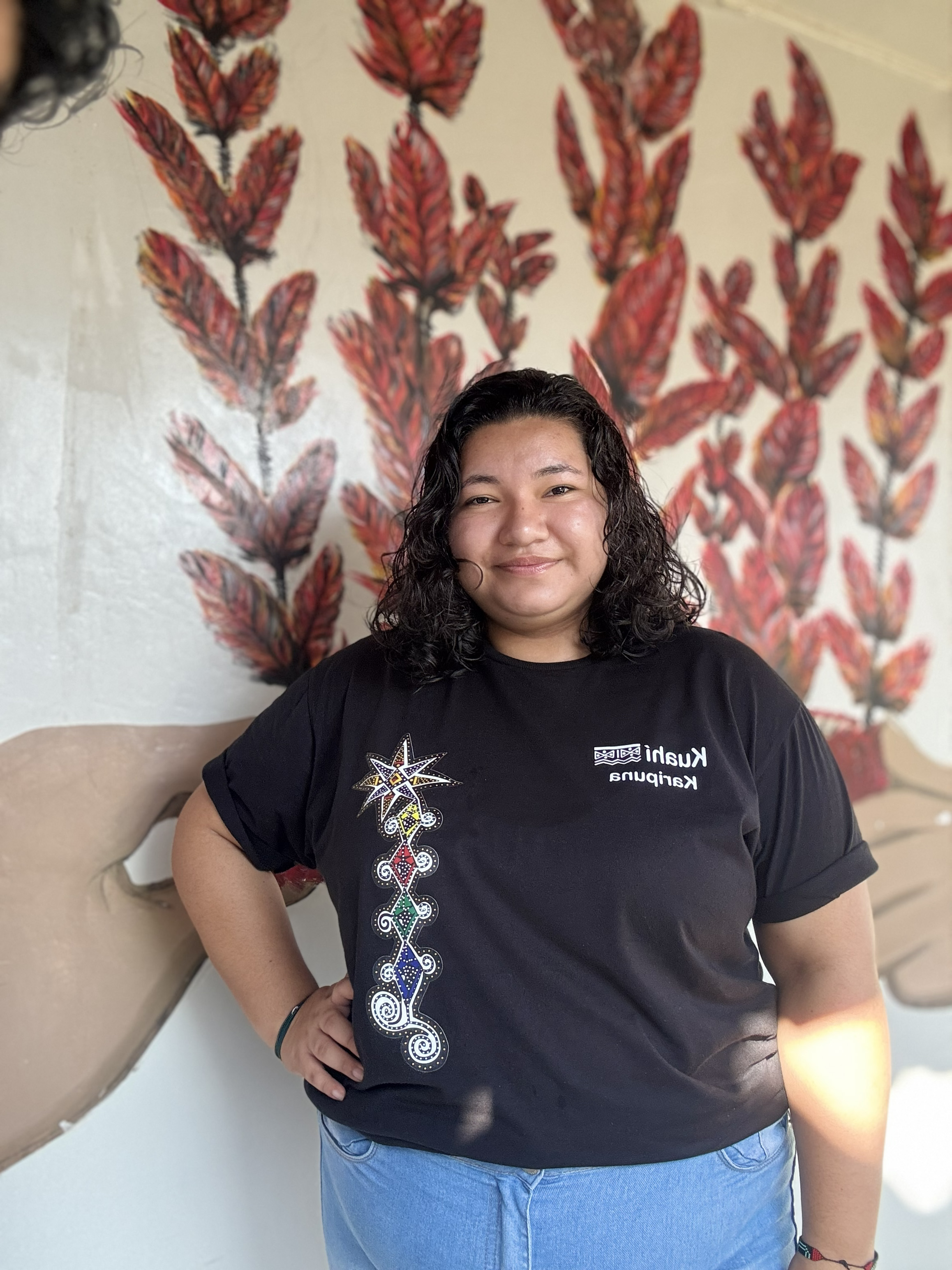 Portrait of Joyce Anika, wearing a black t-shirt and jeans, against a painted wall. 