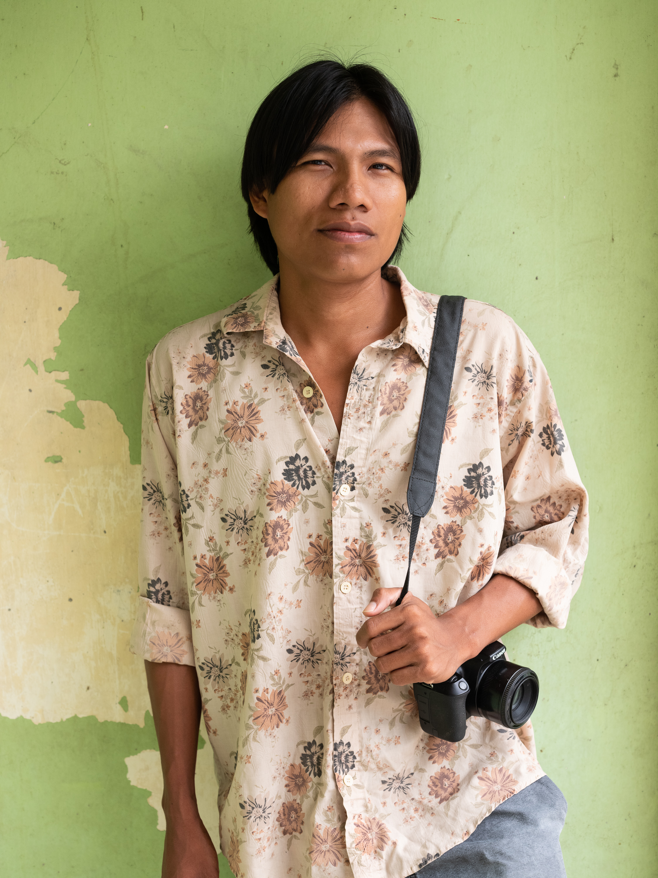 Portrait of Patrick Murayari, wearing jeans and a shirt with a light stamp, holding his camera on his shoulder against a pale green wall. 