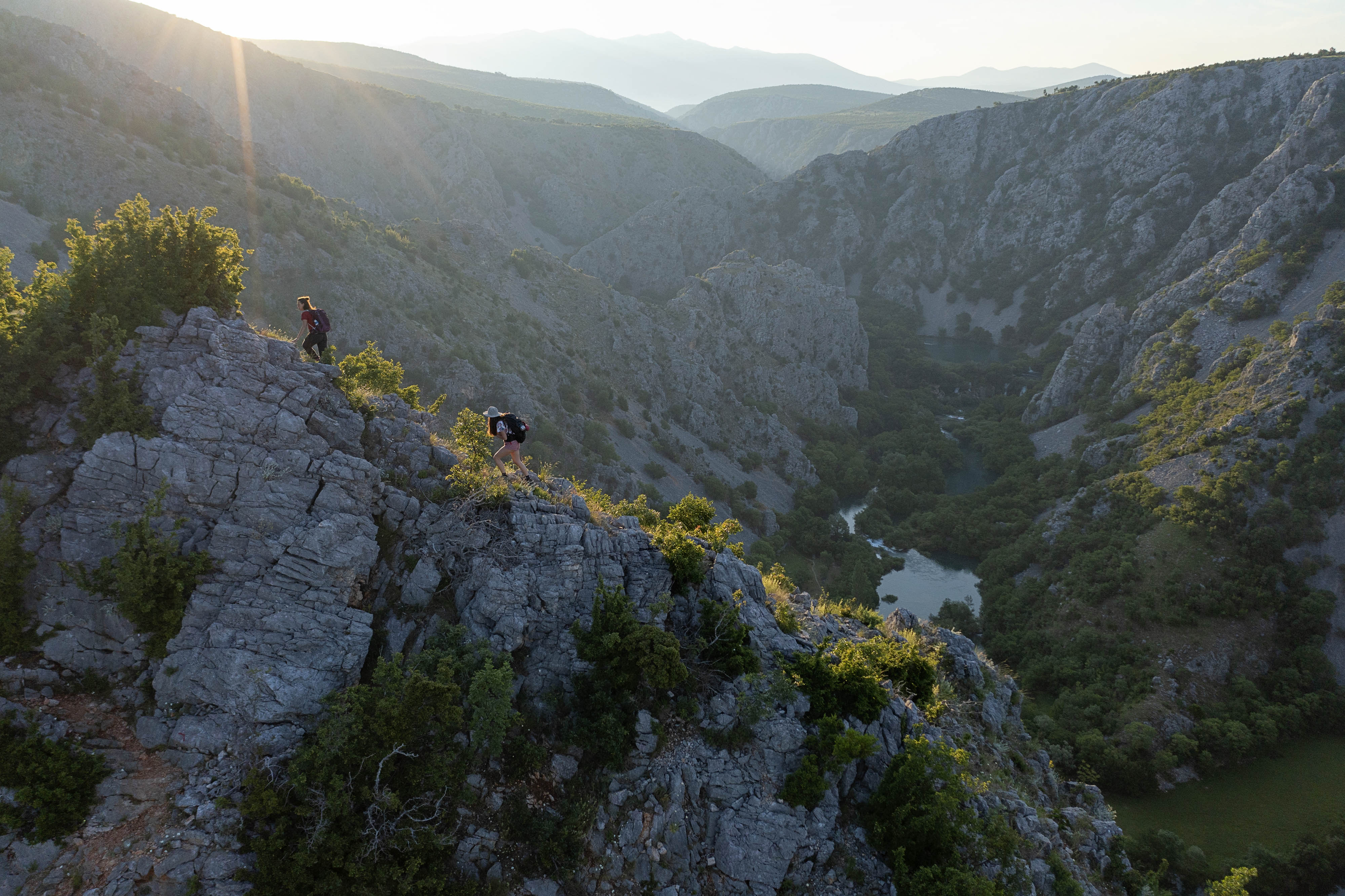 Hikers scale a steep ridge of rock overlooking a river valley.