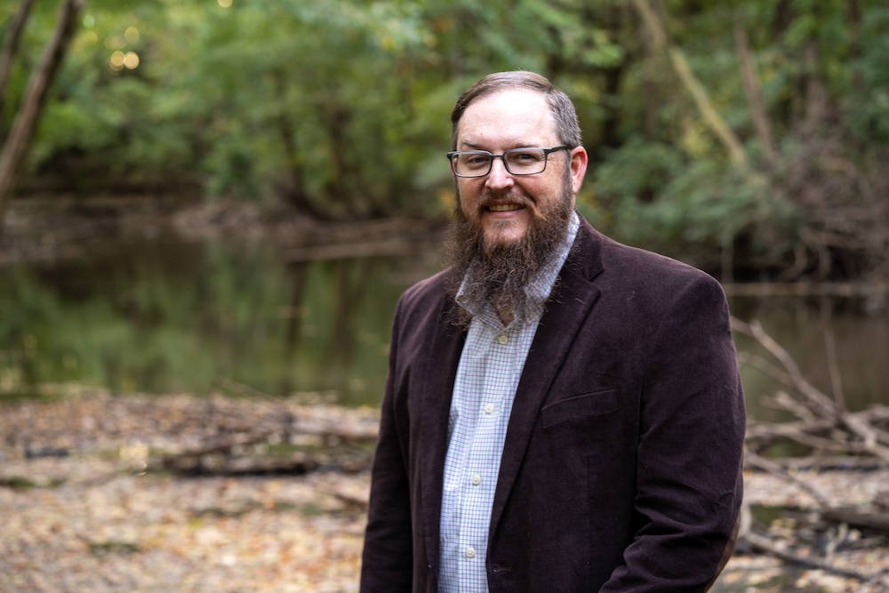 Man with a brown blazer and a blue striped shirt posing in front of a body of water.
