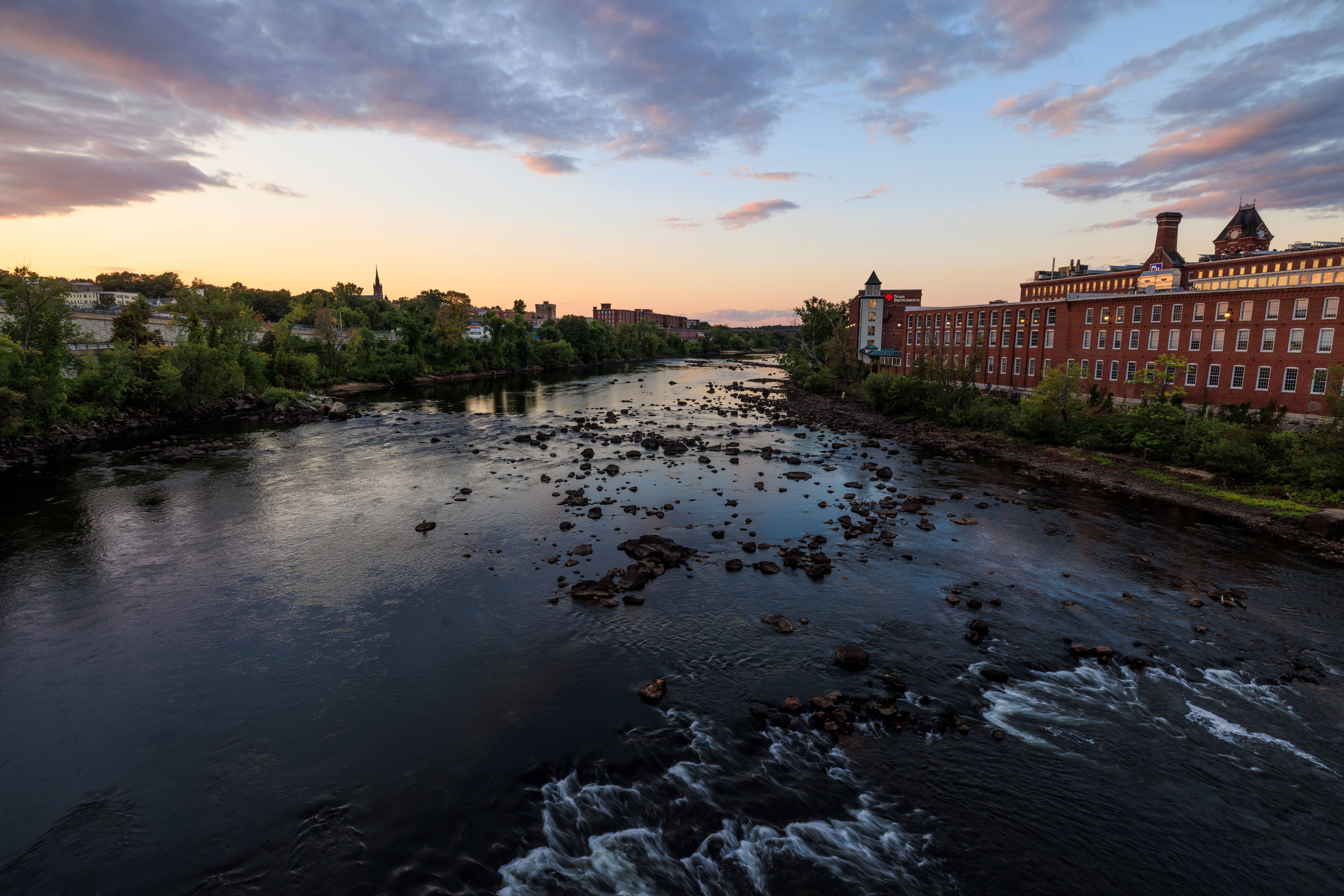 Looking up a river as it flows by a brick mill building. The sun is setting in the background.