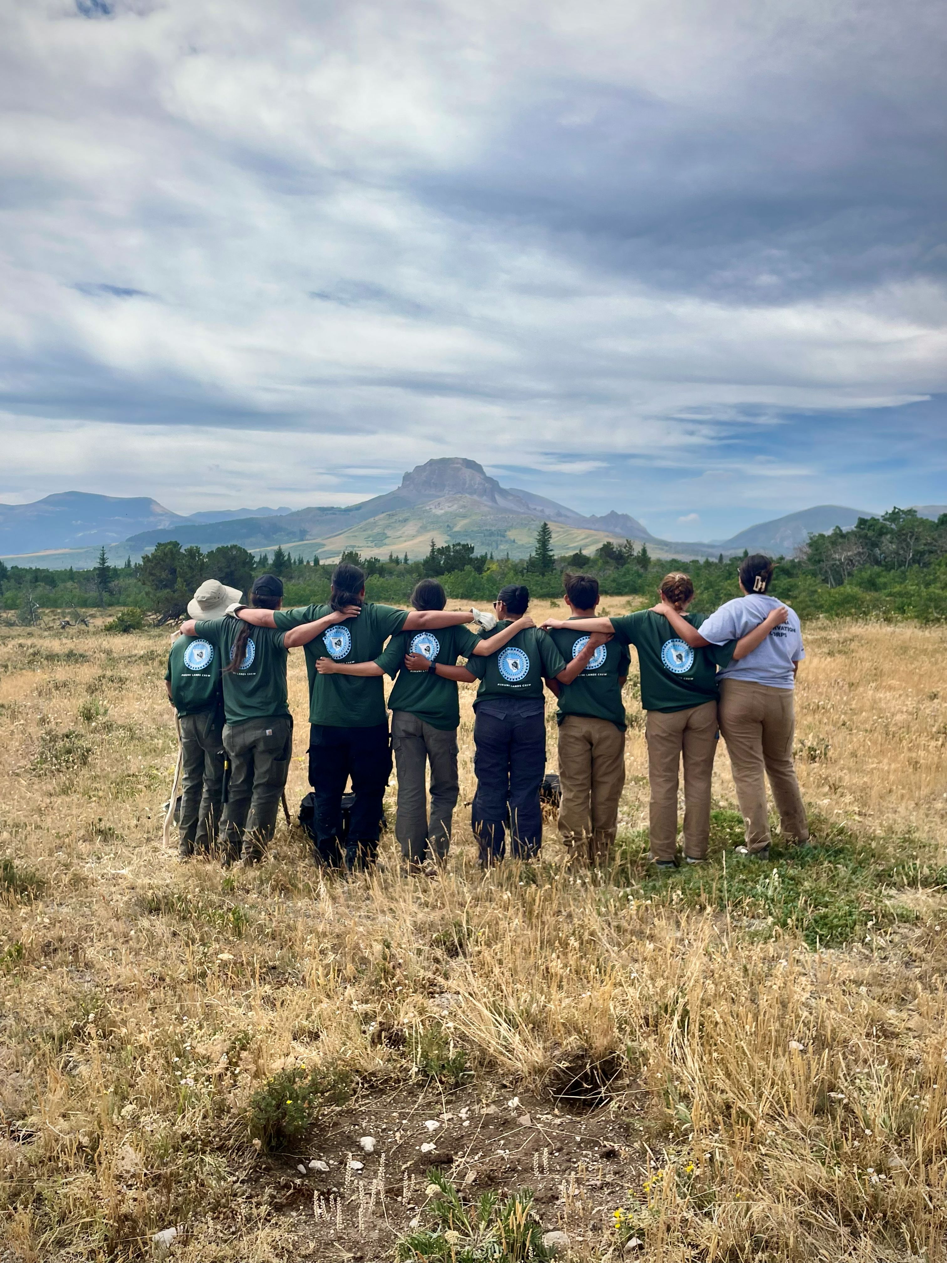 Eight people in matching shirts have hands wrapped around each others shoulders as they look into the distance.