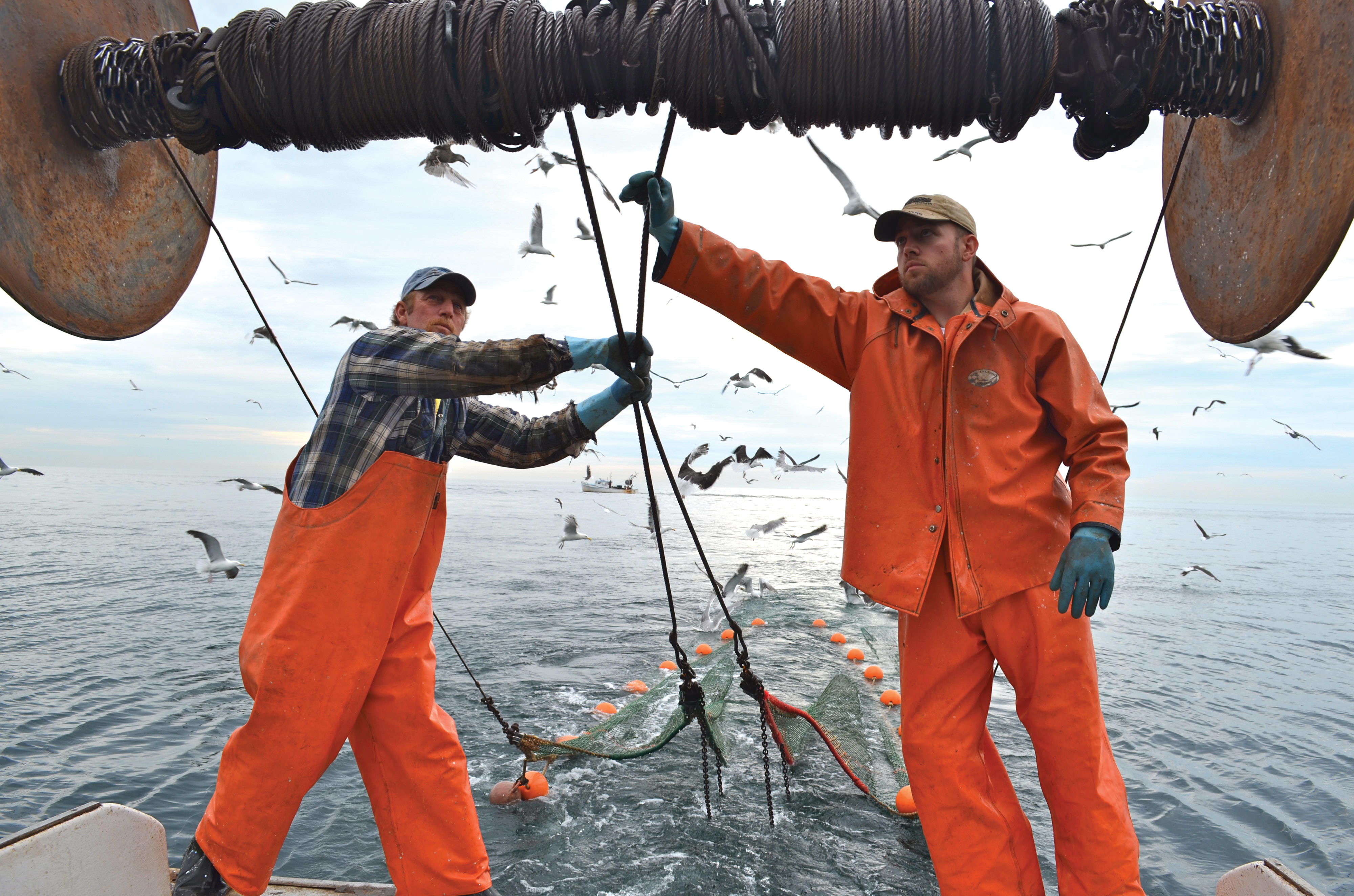 Two fishermen in bright orange jackets pull rigging on a fishing boat.