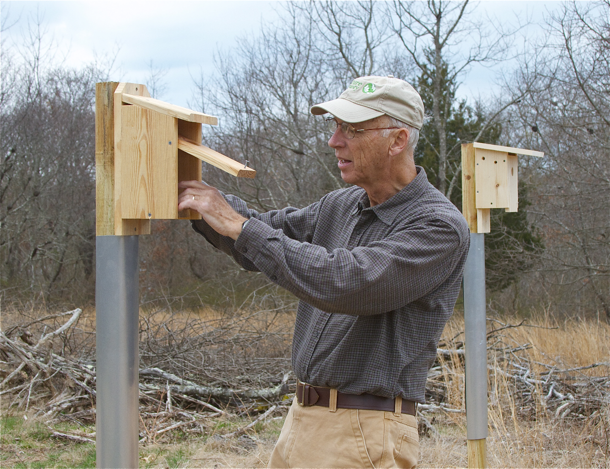 A man wearing a hat opens the door to a wooden bird house.