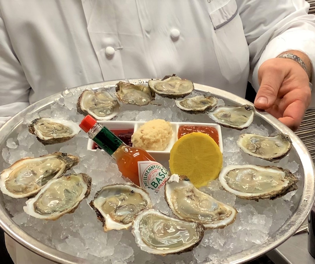A dozen oysters and a bottle of Tabasco are presented on a silver tray.