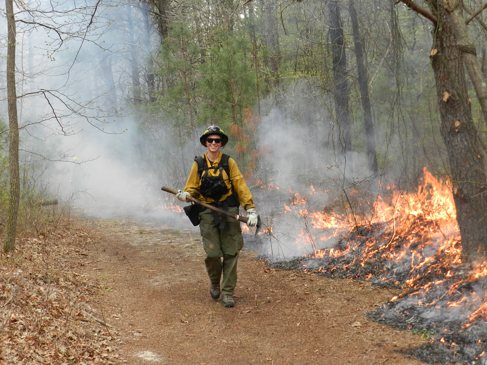 A photo of Natasha Whetzel walking on a path in the forest wearing yellow fire gear and holding an axe as a small flame burns to her right.