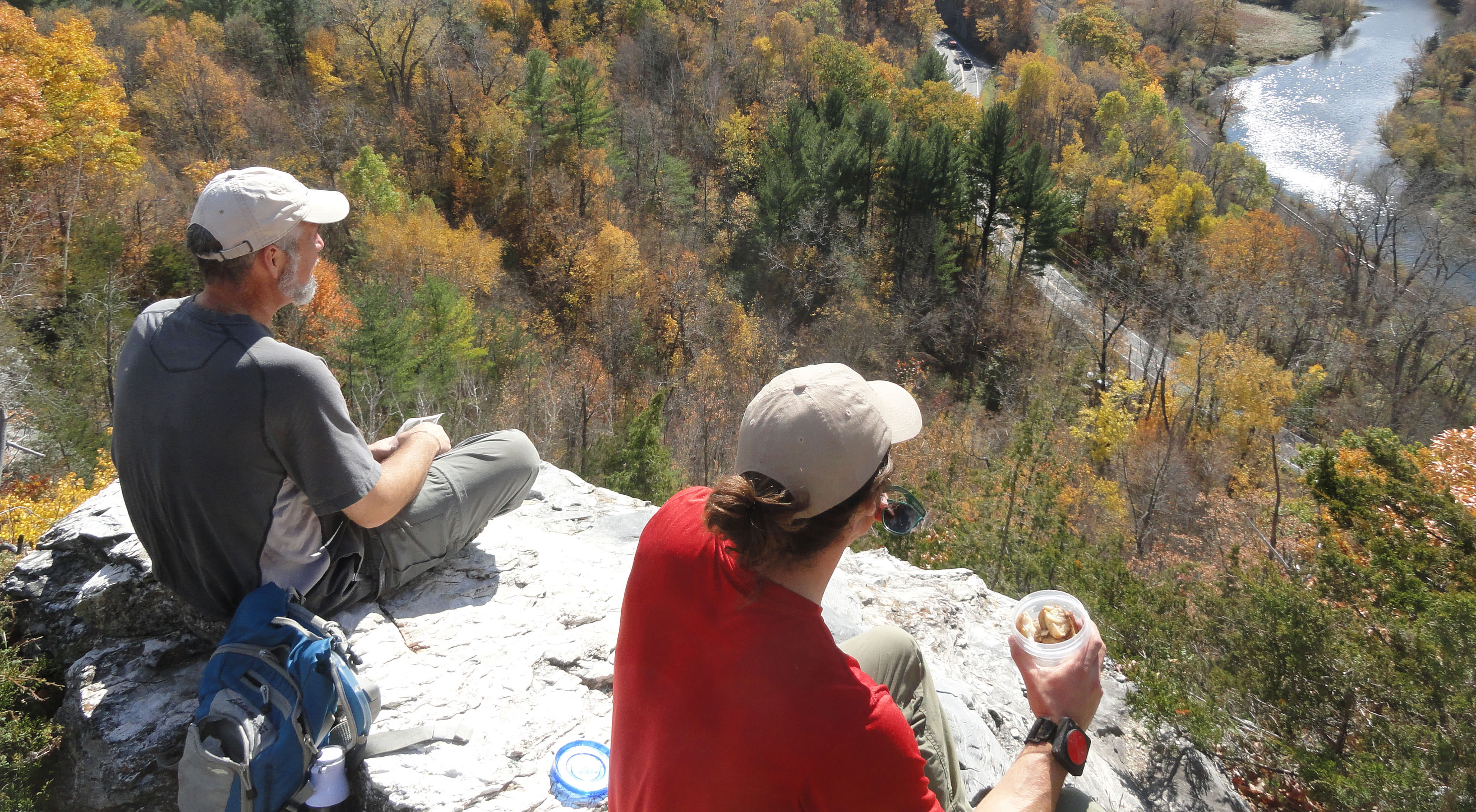 Two people sit on a rock ledge high above a forest with a river running through it.