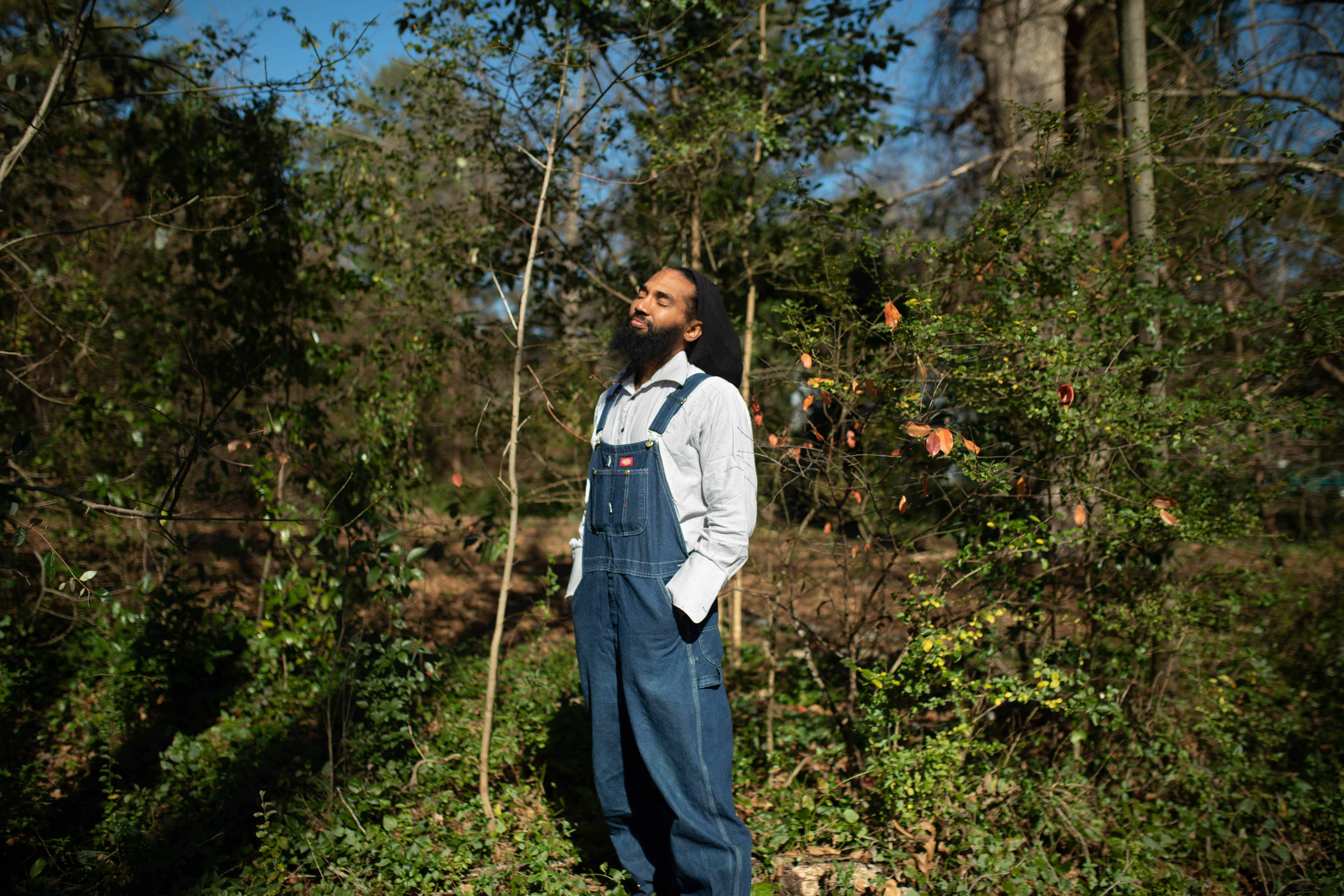A man in coveralls with eyes closed stands in an urban garden.