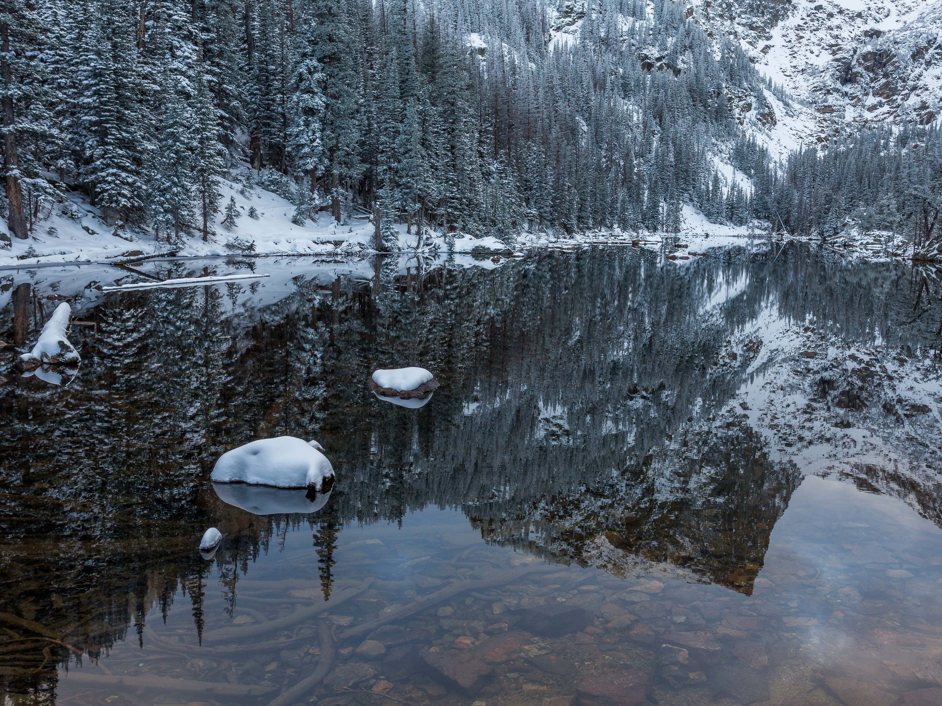 Snow-covered trees surround a lake that reflects Hallet Peak during an early season snowfall in Rocky Mountain National Park.