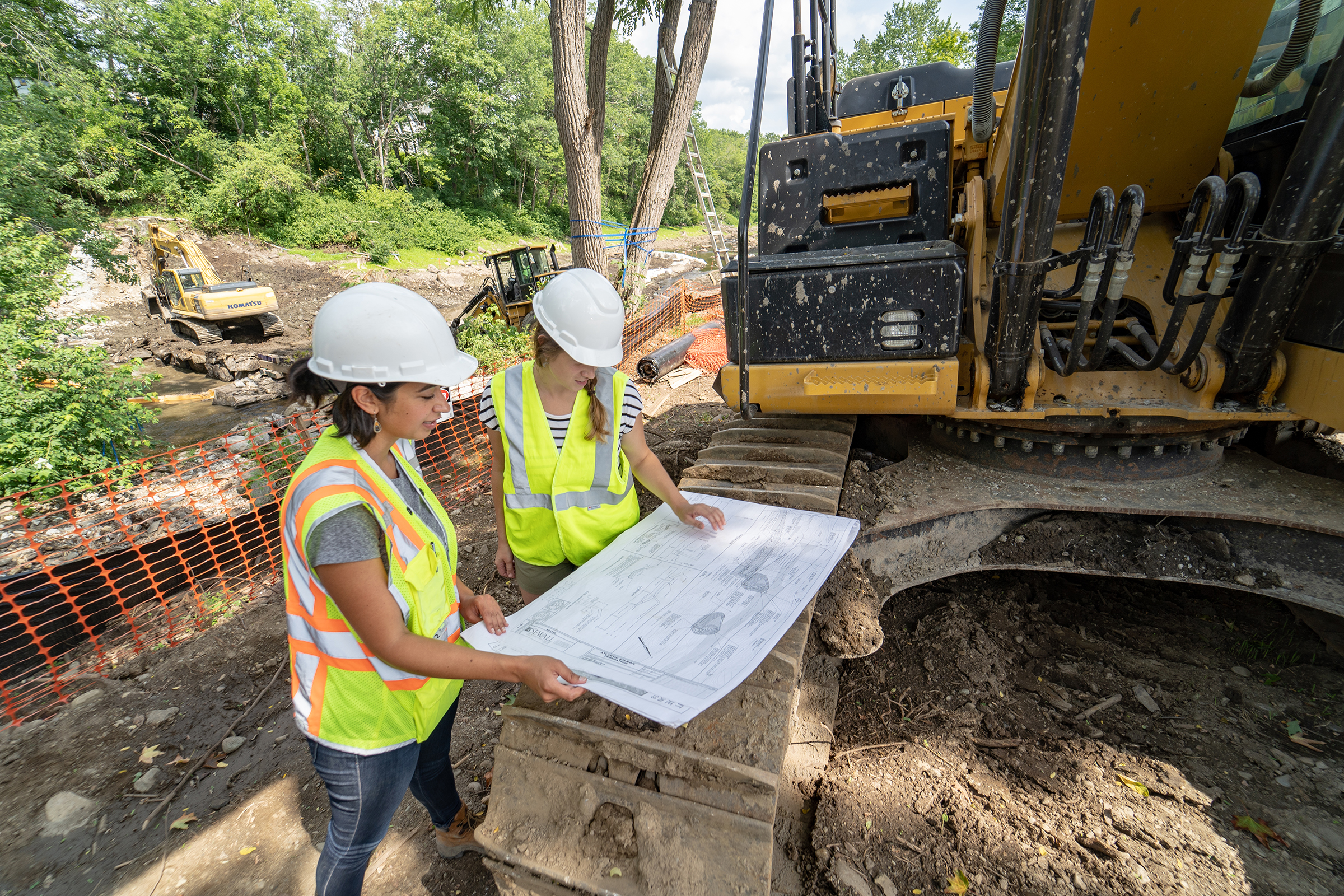 Two people stand next to a piece of heavy construction equipment, studying the map that is spread open on one of its treads.