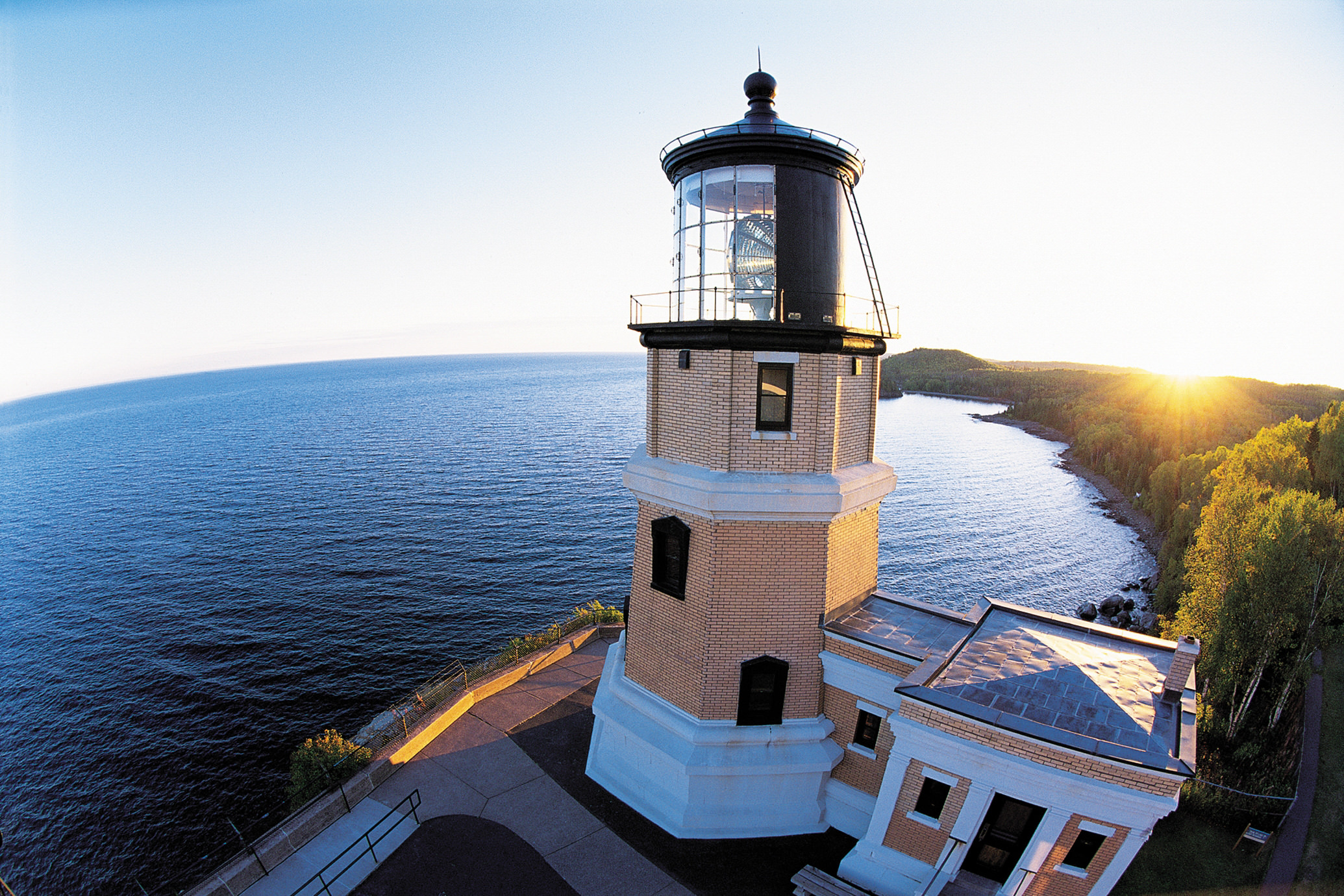 Lighthouse in foreground with vast lake and sun setting over rolling forested land in the background.