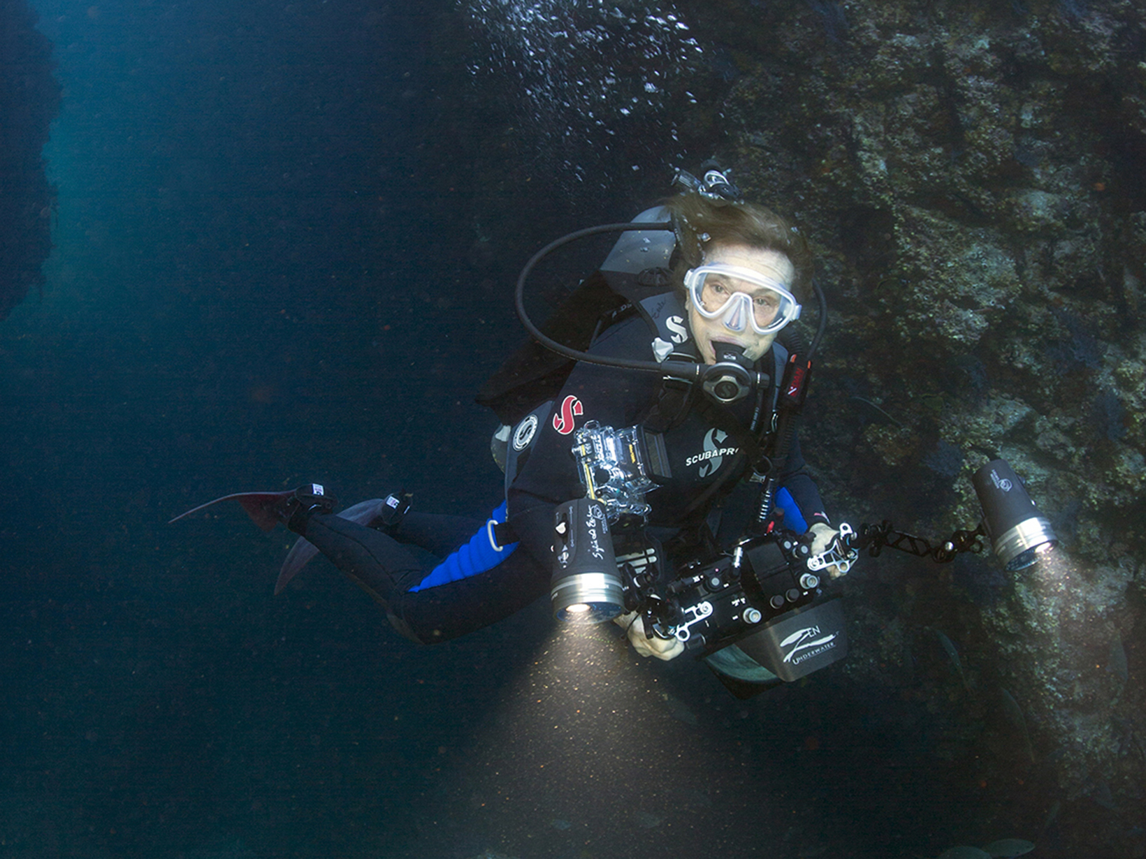 A woman in a dive suit, mask and camera explores a sea cave. 
