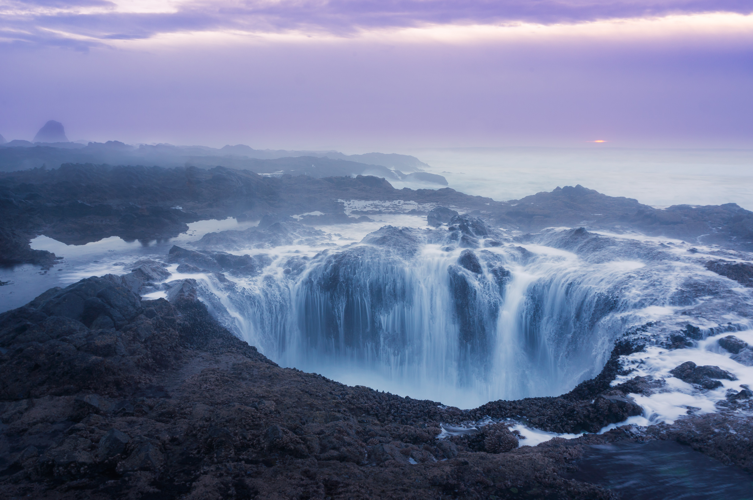 Tidal waters rushing into a large hole in black coastal rocks surrounded by blue sea and purple skies.