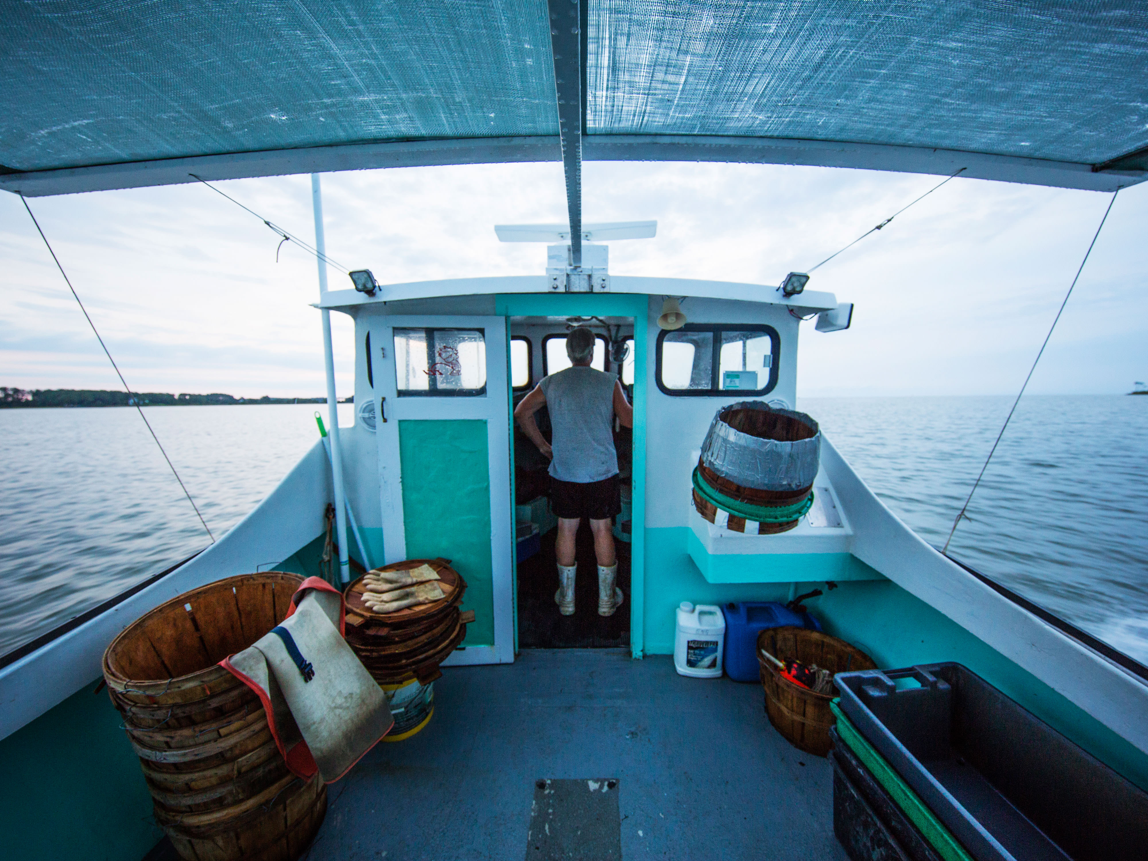 A man stand in the wheelhouse of a small boat. His back is to the camera. Large bushel baskets are stacked behind him on the deck. The still water of the Chesapeake Bay stretches out to the horizon.