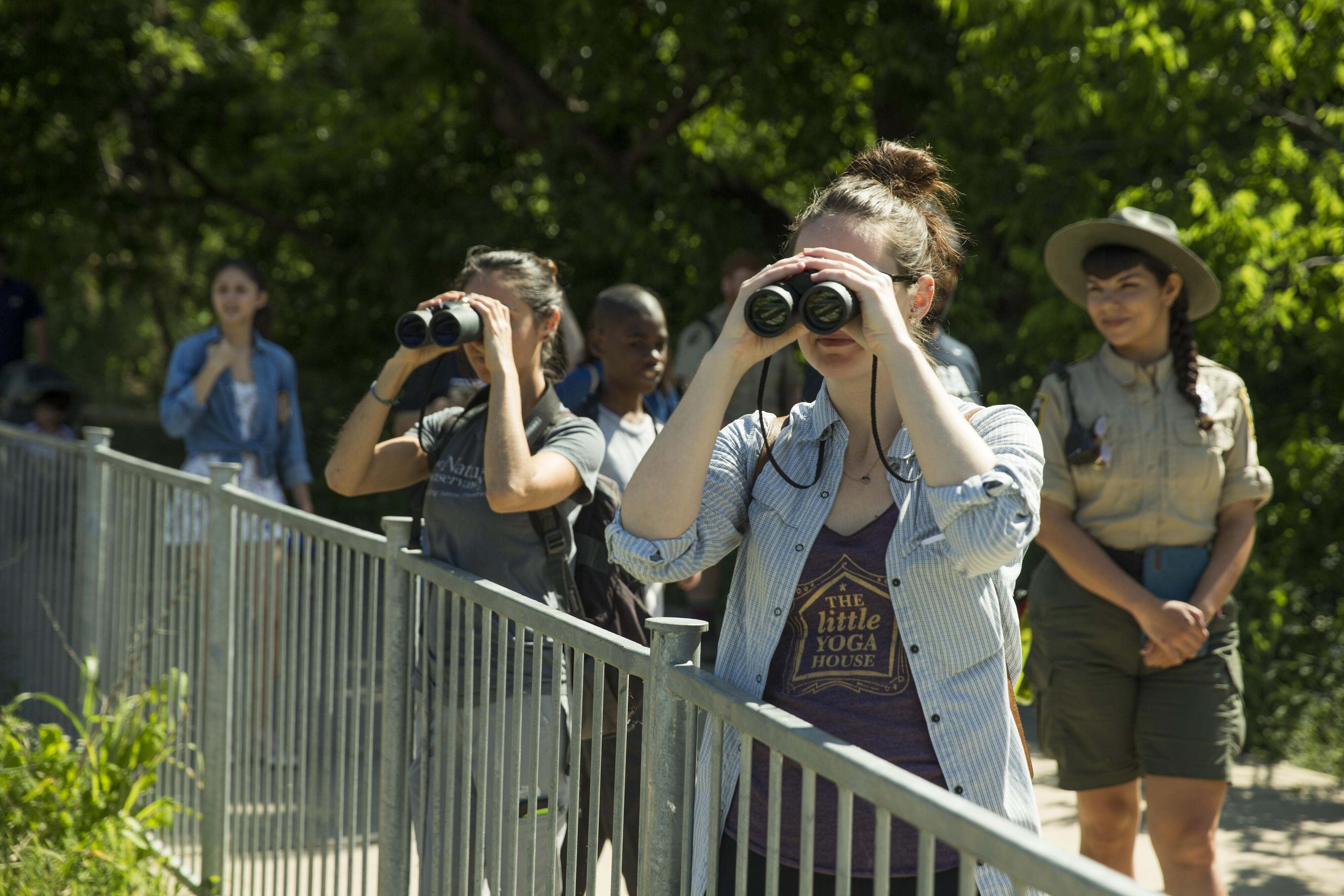Two people stand together behind a fence, each holding a set of binoculars to their eyes.