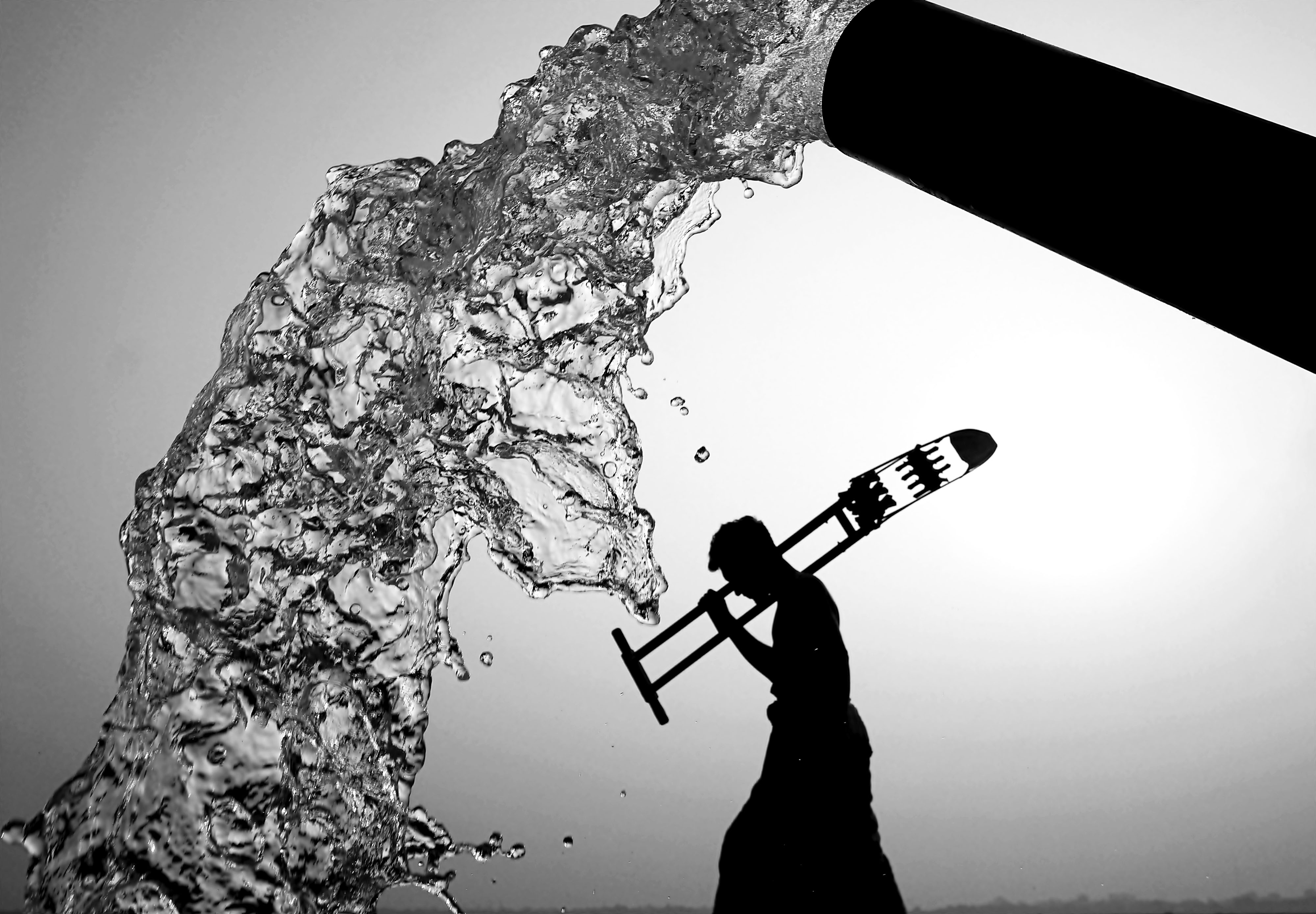 A black and white view of water pouring from a spout and the silhouette of a person walking and carrying a large tool.