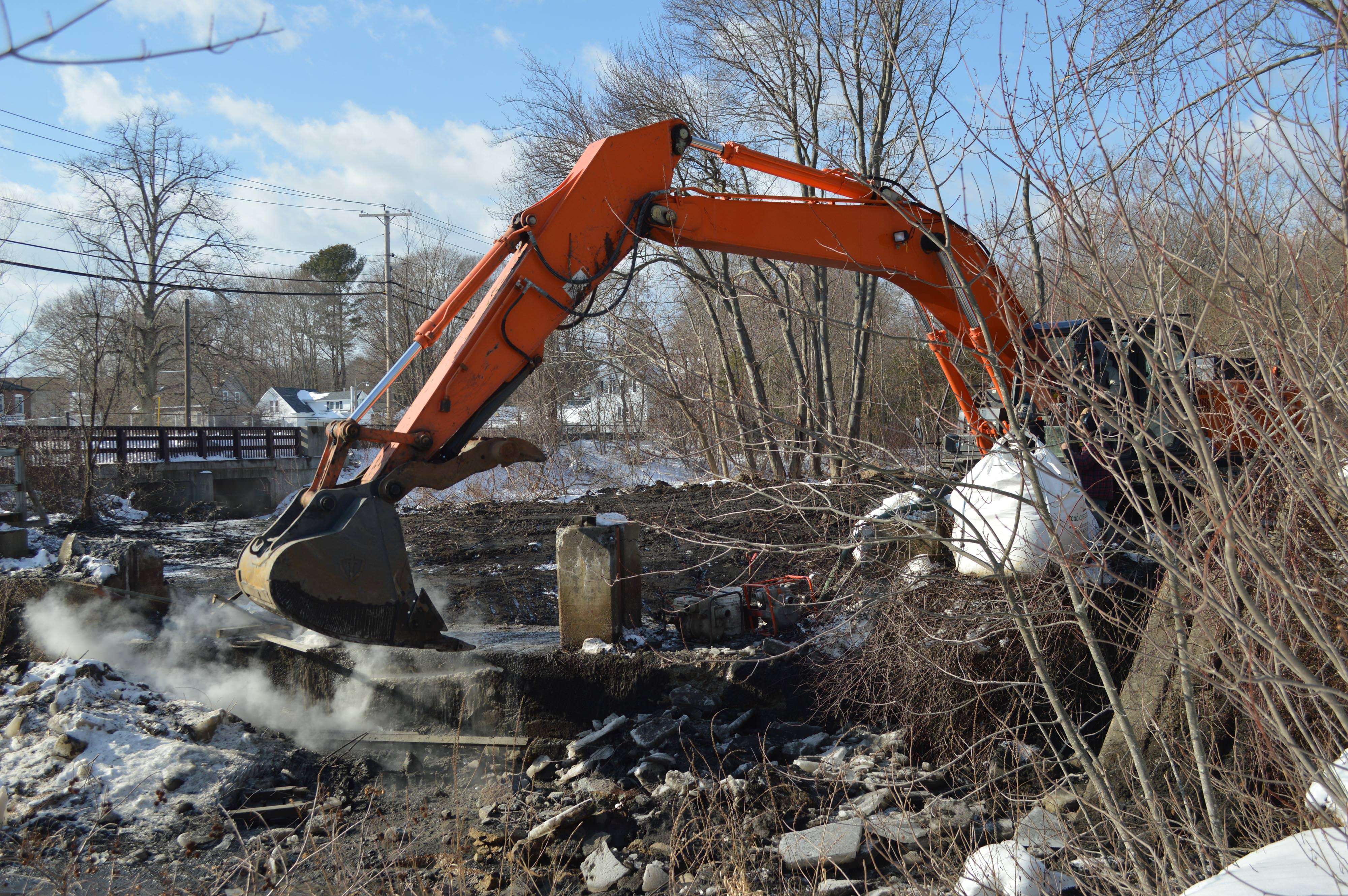 An orange excavator arm poised above the concrete remains of the West Britannia Dam on a winter day.